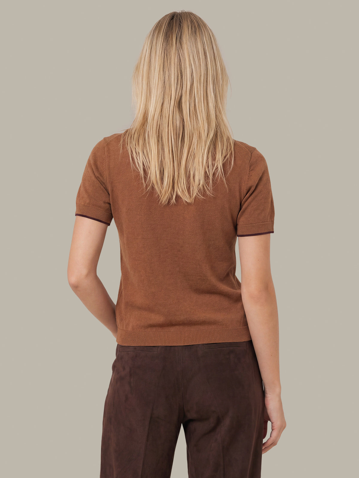 A woman viewed from behind wearing a short-sleeve brown knit top with dark trim and dark brown pants, against a solid light gray background.