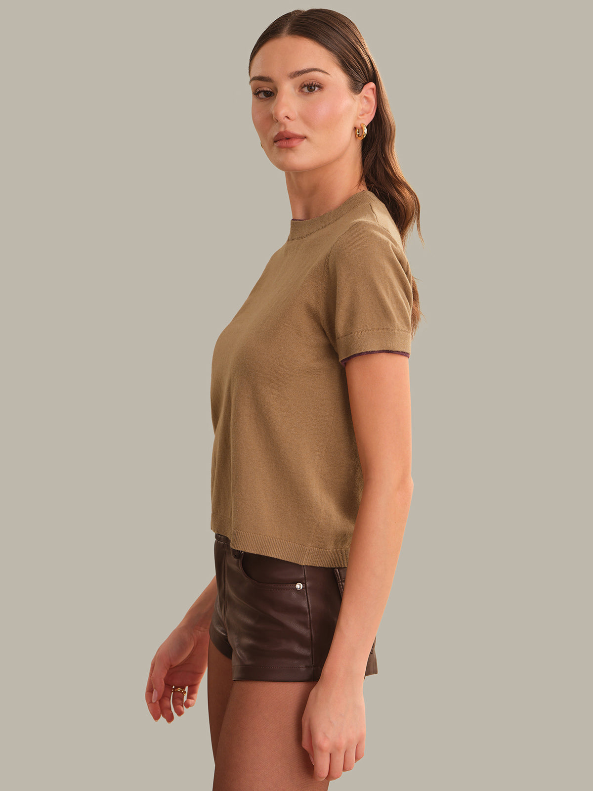 A woman modeling a short-sleeve crewneck t-shirt in a warm tan color, paired with dark brown faux-leather shorts, against a solid light gray background.