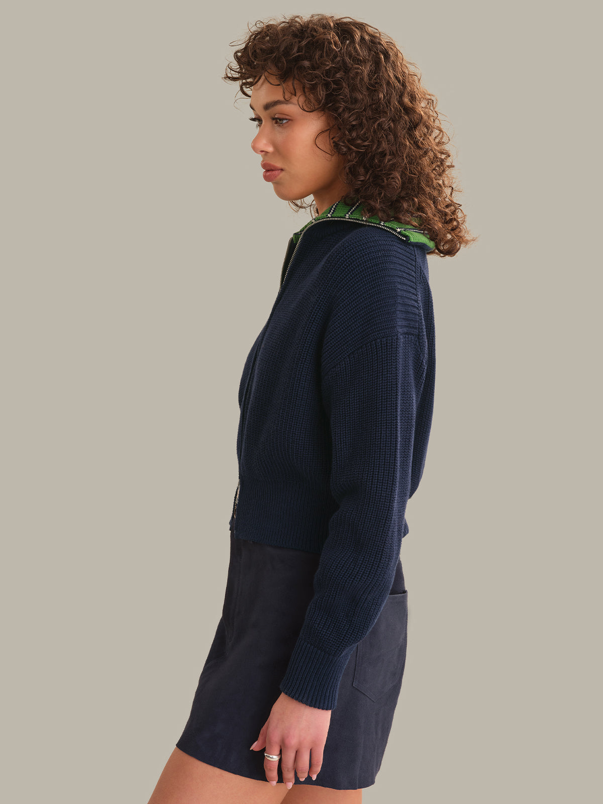 A woman with curly brown hair is shown in profile wearing a dark navy ribbed knit sweater with a green patterned collar, paired with a matching dark skirt, against a solid light gray background.
