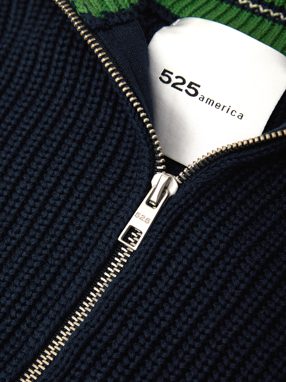Close-up of a navy blue ribbed knit zip-up sweater with a white interior collar label reading '525 america' and a silver zipper.