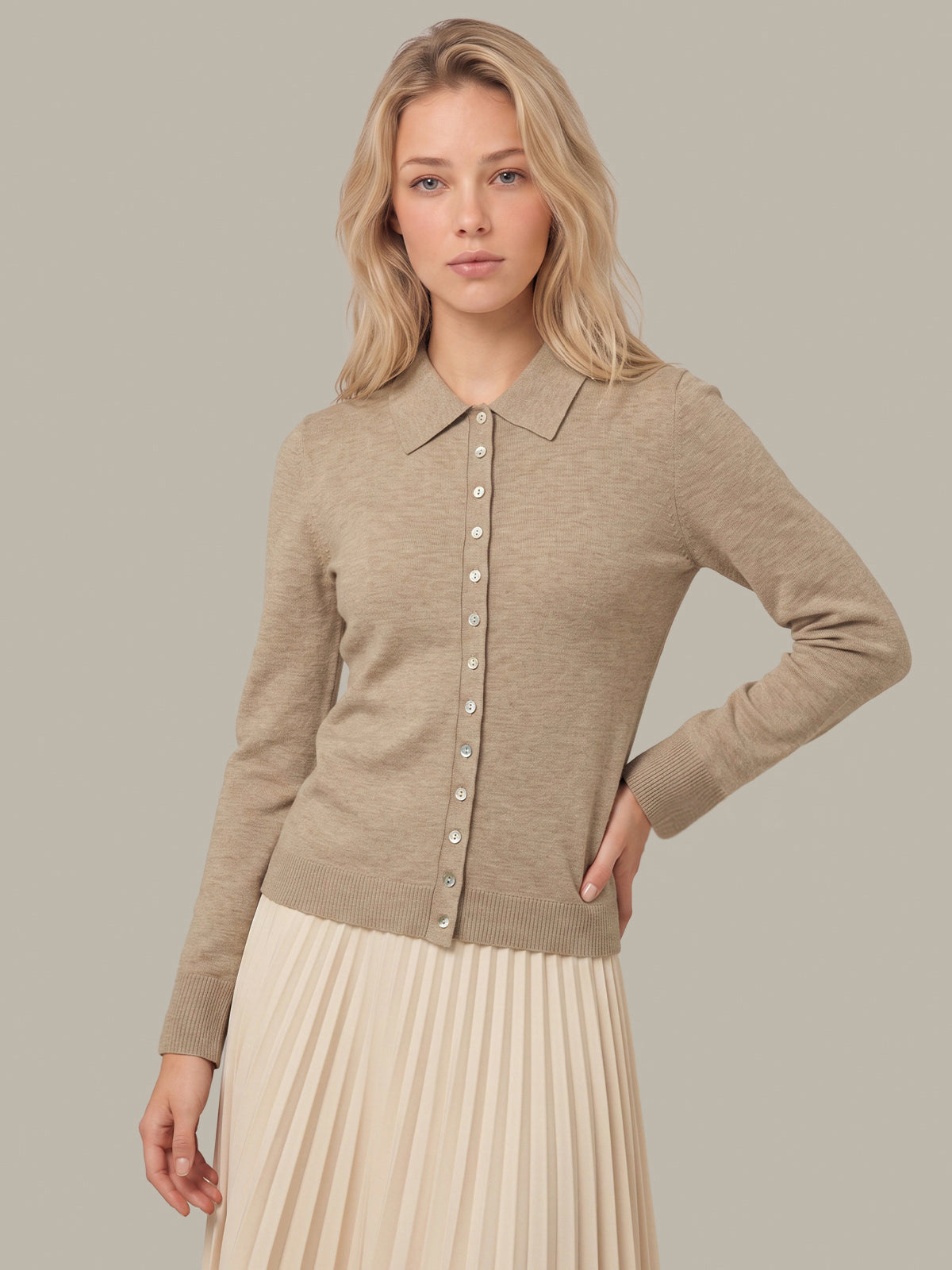 A woman modeling a light beige knit button-front cardigan with a collar, paired with a cream pleated skirt, against a solid taupe background.