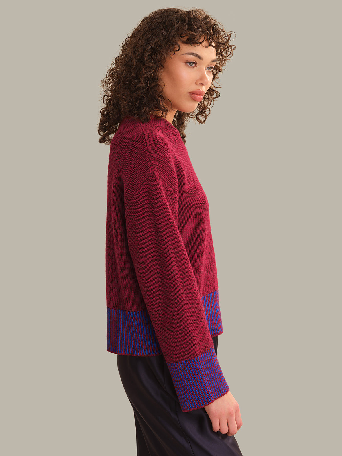 A woman with curly brown hair is shown in profile wearing a ribbed knit sweater in deep burgundy with purple and blue striped ribbed cuffs, paired with dark pants, against a solid light gray background.
