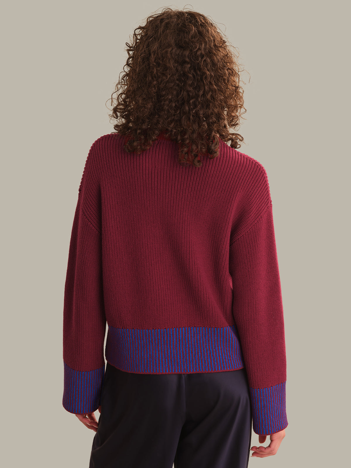 A model viewed from the back wearing a burgundy ribbed knit sweater with blue and red striped cuffs and hem, paired with dark trousers, against a solid light gray background.
