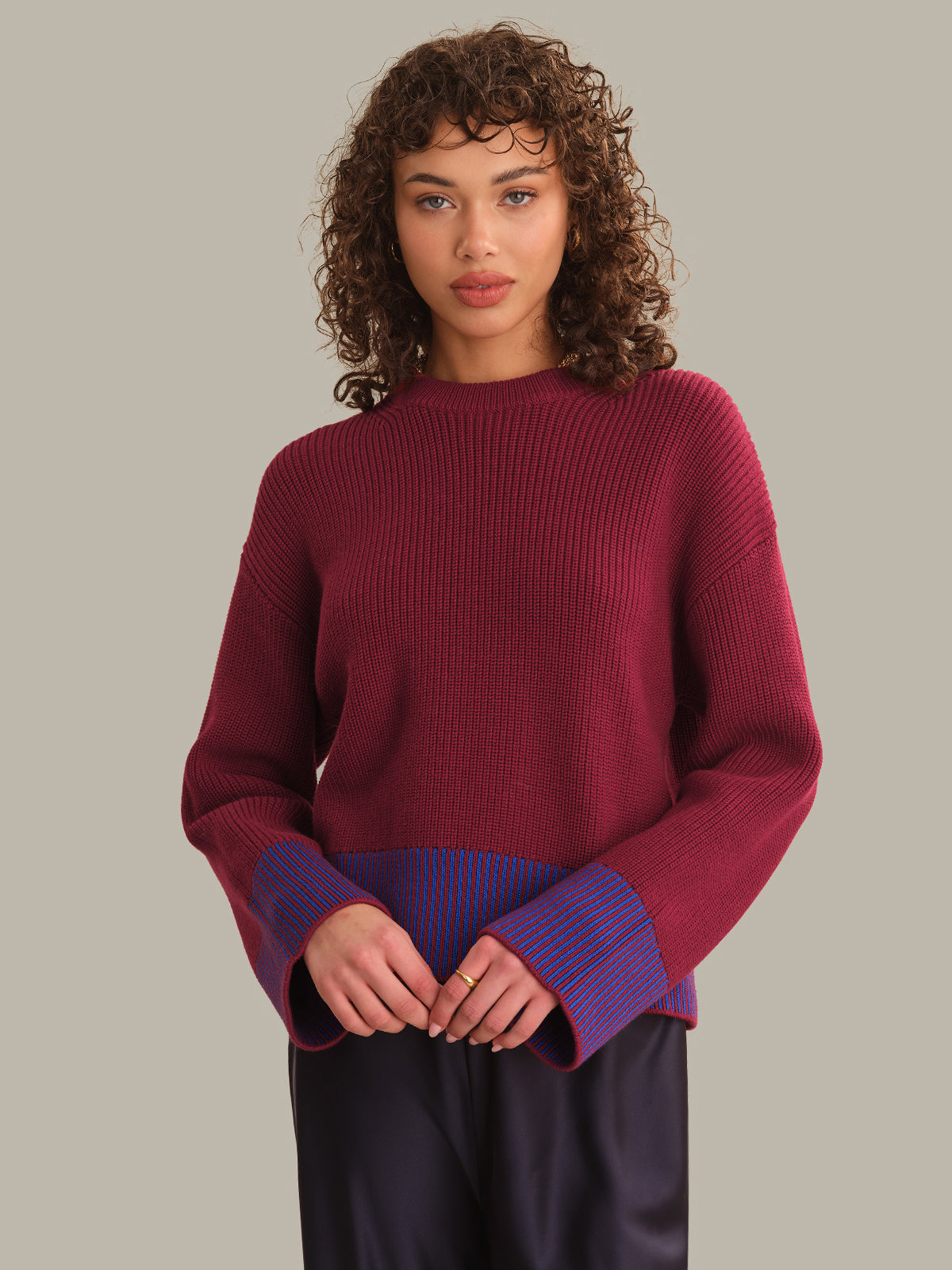 A model with curly brown hair wearing a ribbed knit sweater in deep burgundy with contrasting purple and blue striped cuffs, paired with dark pants, against a solid light gray background.