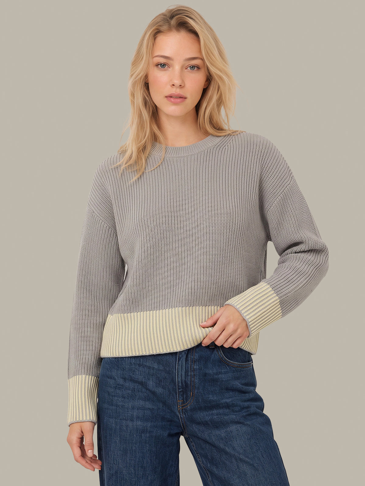A woman modeling a ribbed knit sweater with a light gray body and contrasting cream ribbed cuffs and hem, paired with blue jeans against a solid light gray background.