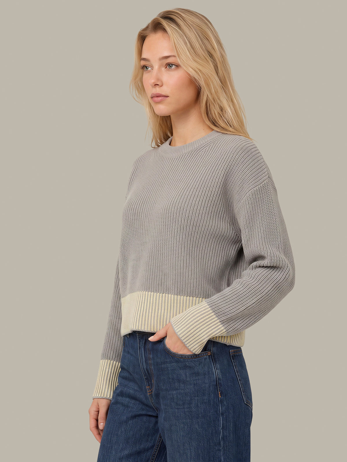 A woman wearing a ribbed knit sweater with a light gray body and cream-colored ribbed trim at the hem and cuffs, paired with blue jeans, against a solid light gray background.