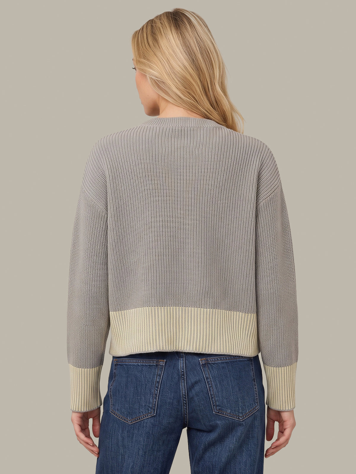 A woman shown from the back wearing a ribbed knit sweater with a light gray body and contrasting cream ribbed bands at the hem and cuffs, paired with blue jeans against a solid taupe background.
