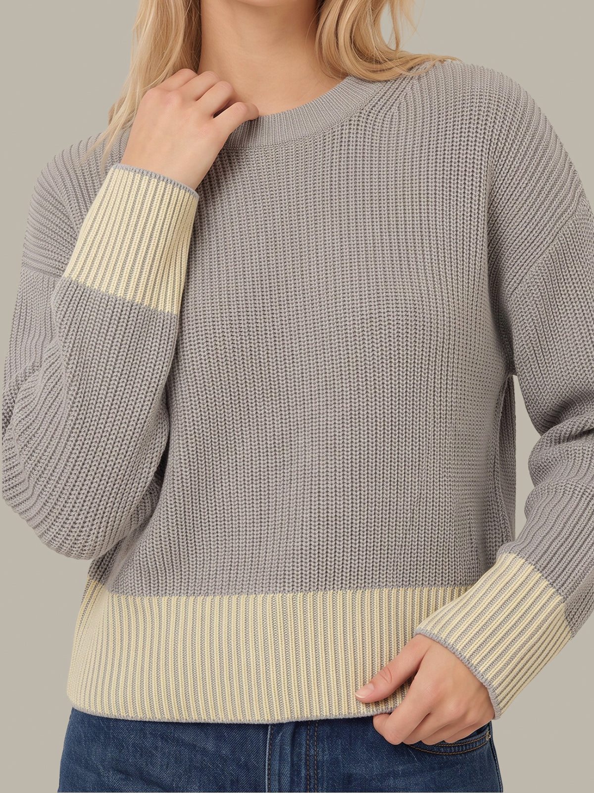 A woman wearing a gray ribbed knit sweater with contrasting cream ribbed cuffs and hem, paired with blue jeans against a neutral studio background.