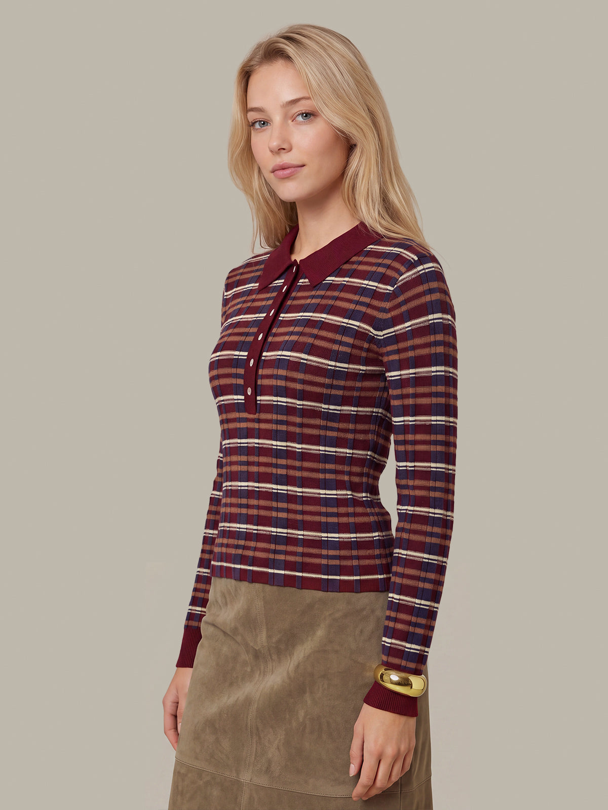 A woman modeling a long-sleeve knit polo shirt with a red, navy, and white plaid pattern and a solid burgundy collar, paired with a brown skirt, against a solid beige background.