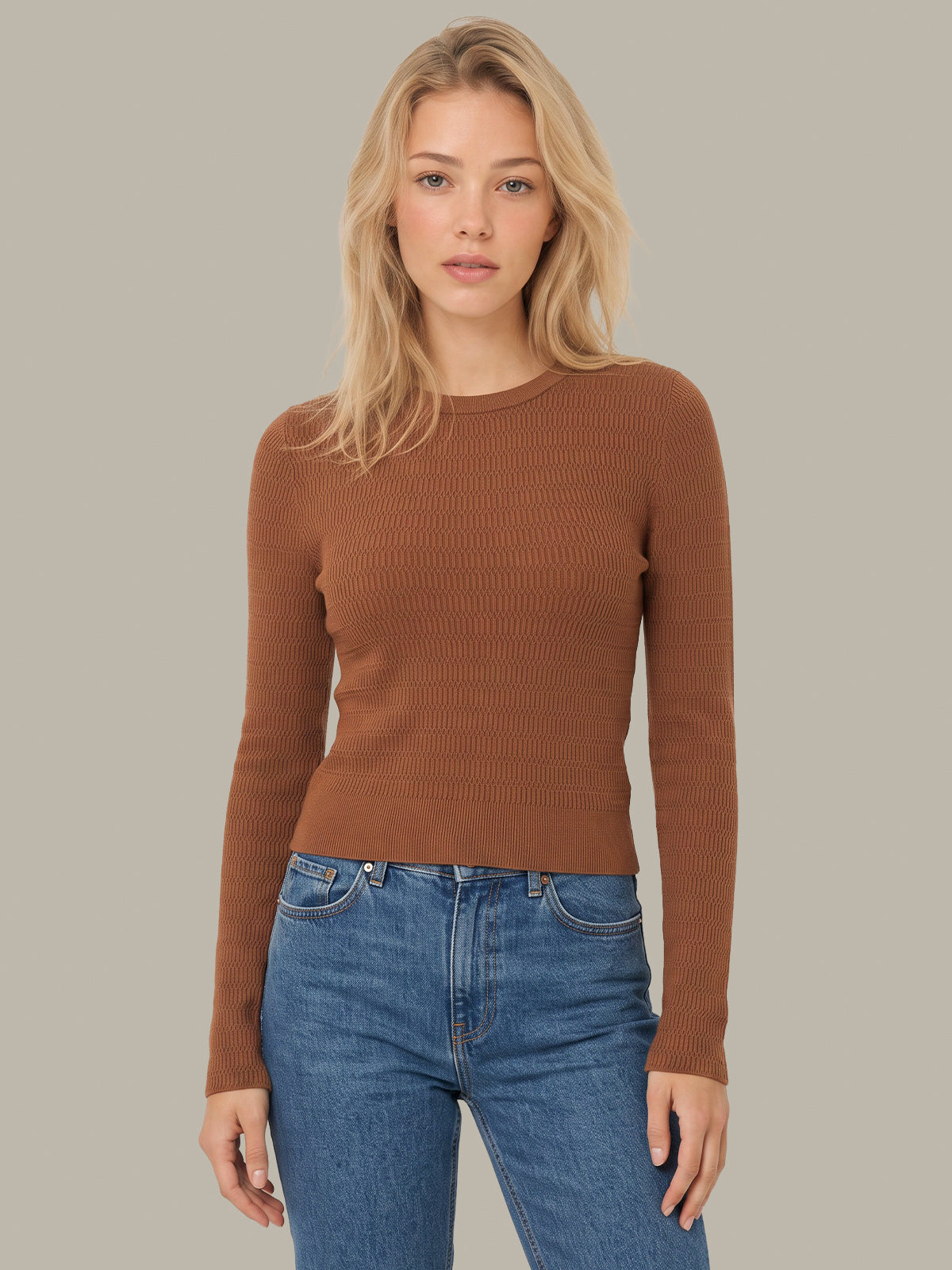 A blonde woman wearing a fitted, long-sleeve ribbed knit sweater in a warm brown/caramel shade, paired with blue jeans, against a solid neutral gray background.