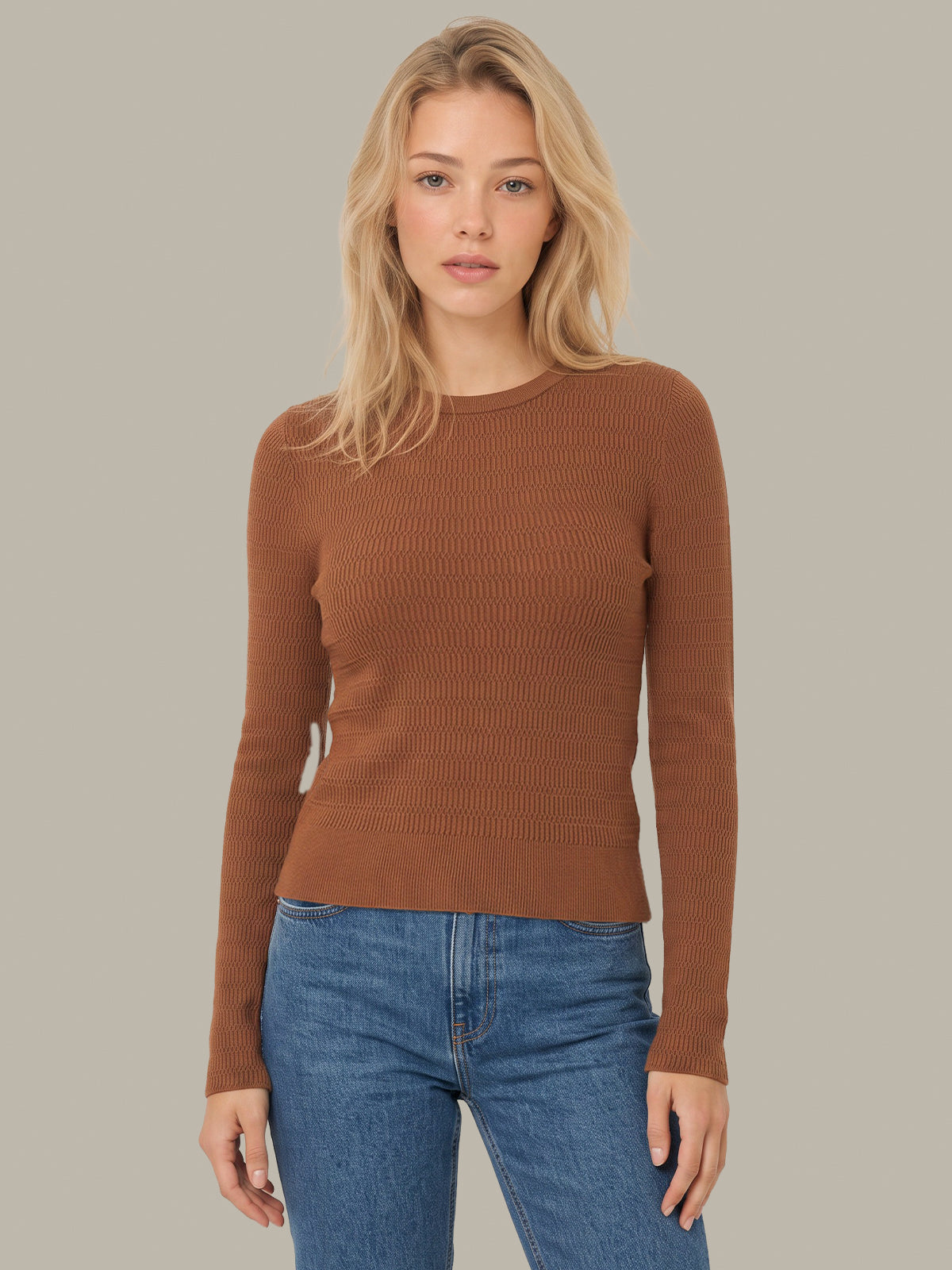 A blonde woman wearing a ribbed long-sleeve crewneck sweater in a warm brown color, paired with blue jeans, against a solid light gray background.