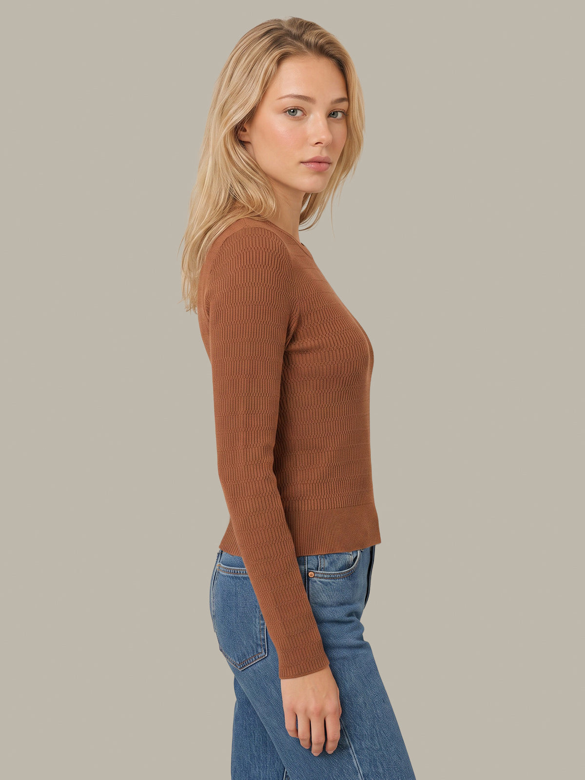 A woman shown in side profile wearing a fitted ribbed long-sleeve knit top in a warm camel brown color, paired with blue jeans, against a solid light gray background.