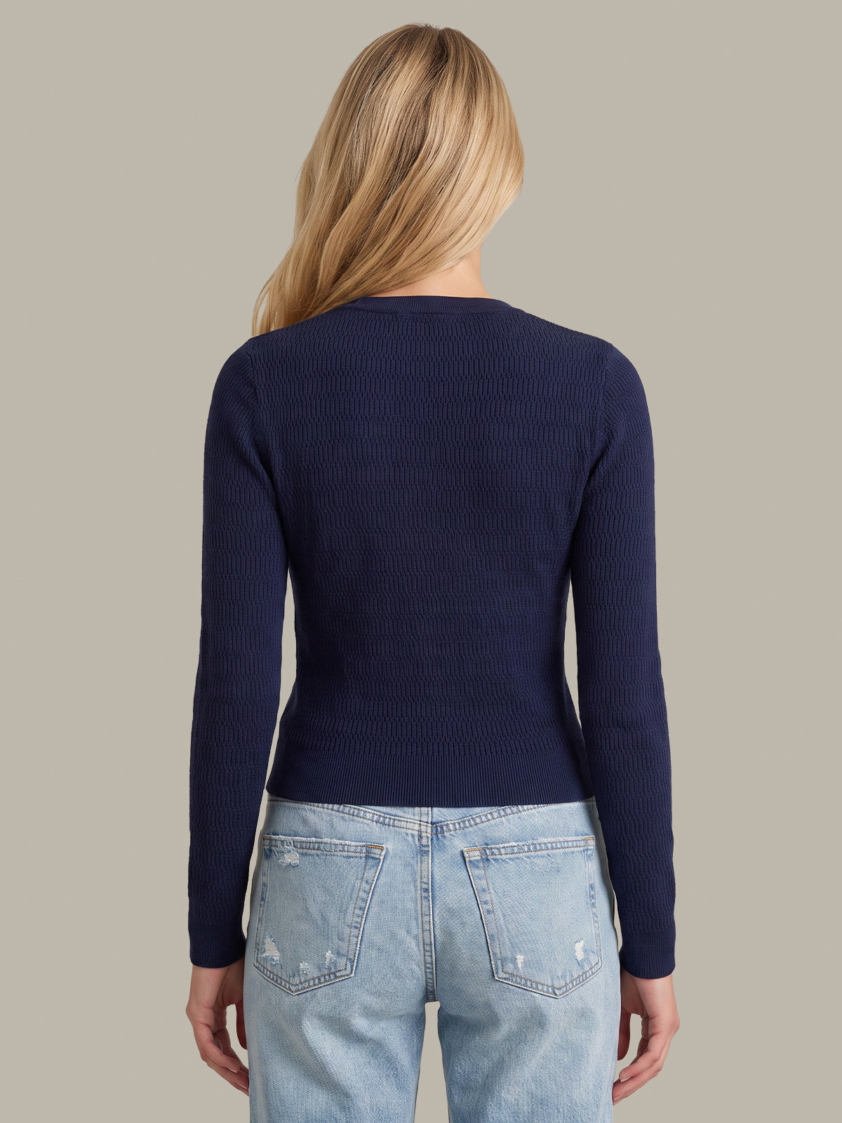 A woman viewed from the back wearing a navy blue ribbed long-sleeve top and light blue jeans, set against a solid taupe background.