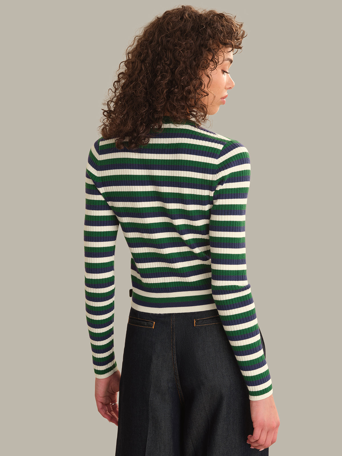 A woman with curly brown hair is shown from the back wearing a green and white striped ribbed knit sweater and dark denim pants, set against a solid light gray background.