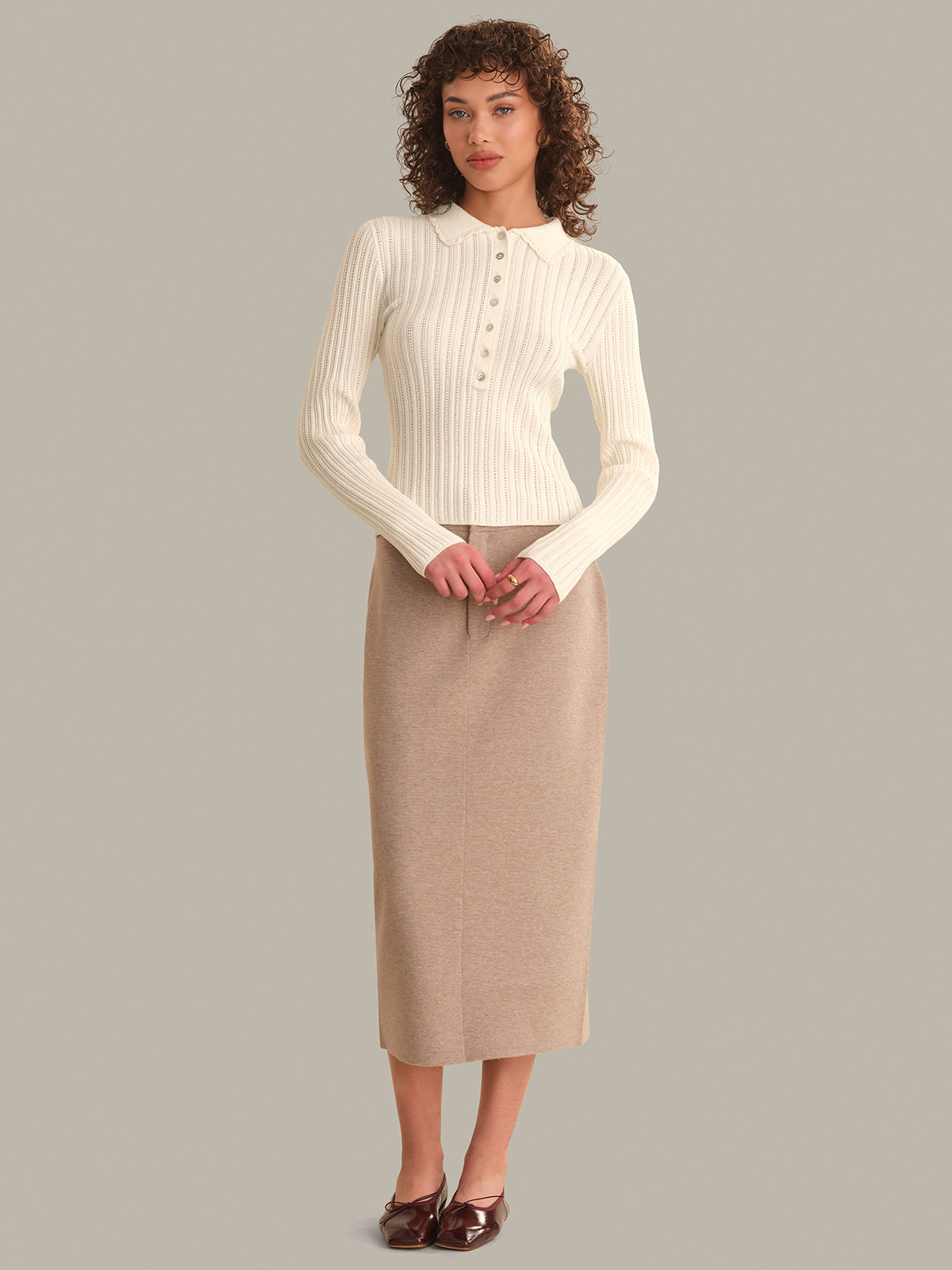 Woman wearing a cream ribbed sweater and beige pencil skirt on a gray background