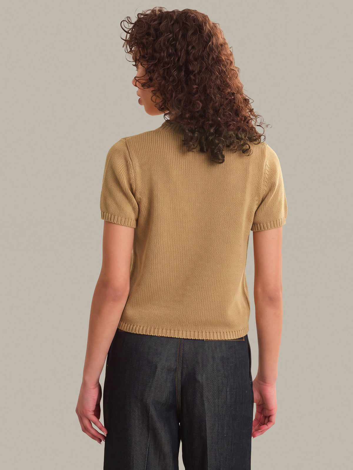 A woman with curly brown hair is shown from the back wearing a short-sleeve ribbed knit top in camel beige and dark charcoal pants, set against a solid light gray studio background.