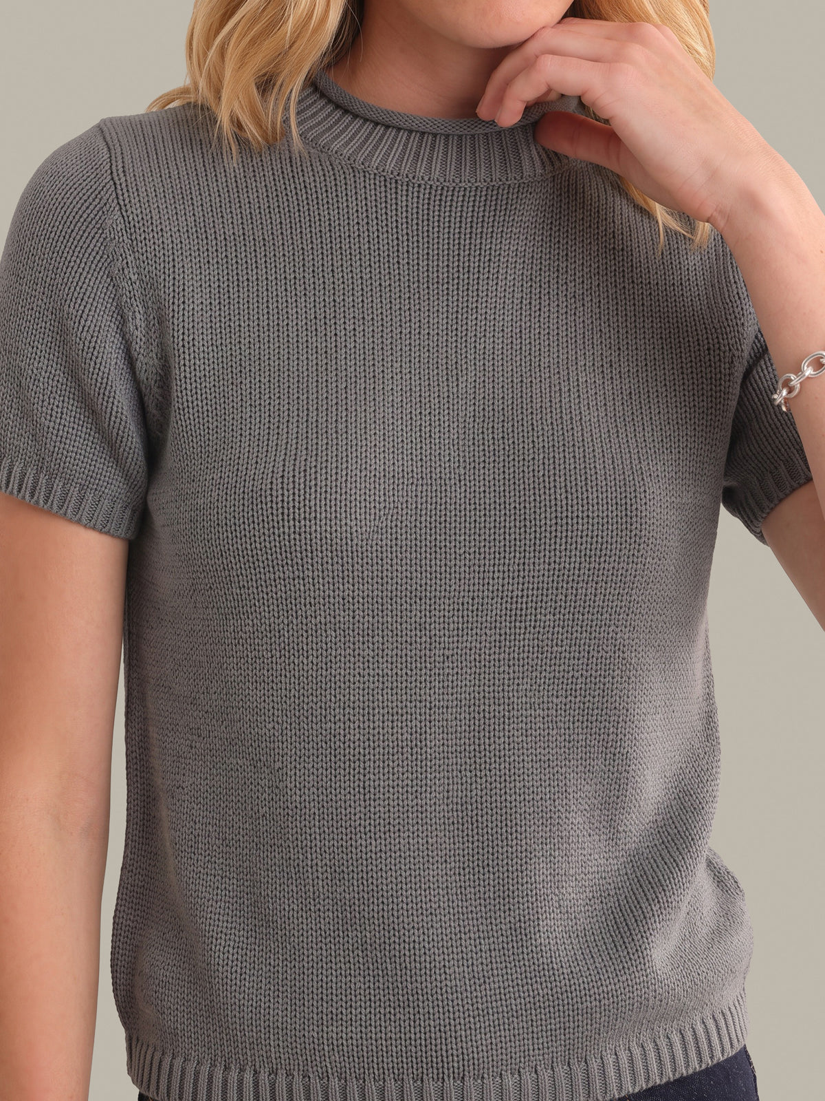 A close-up of a woman wearing a gray short-sleeve ribbed knit sweater with a mock neck, set against a solid neutral background.