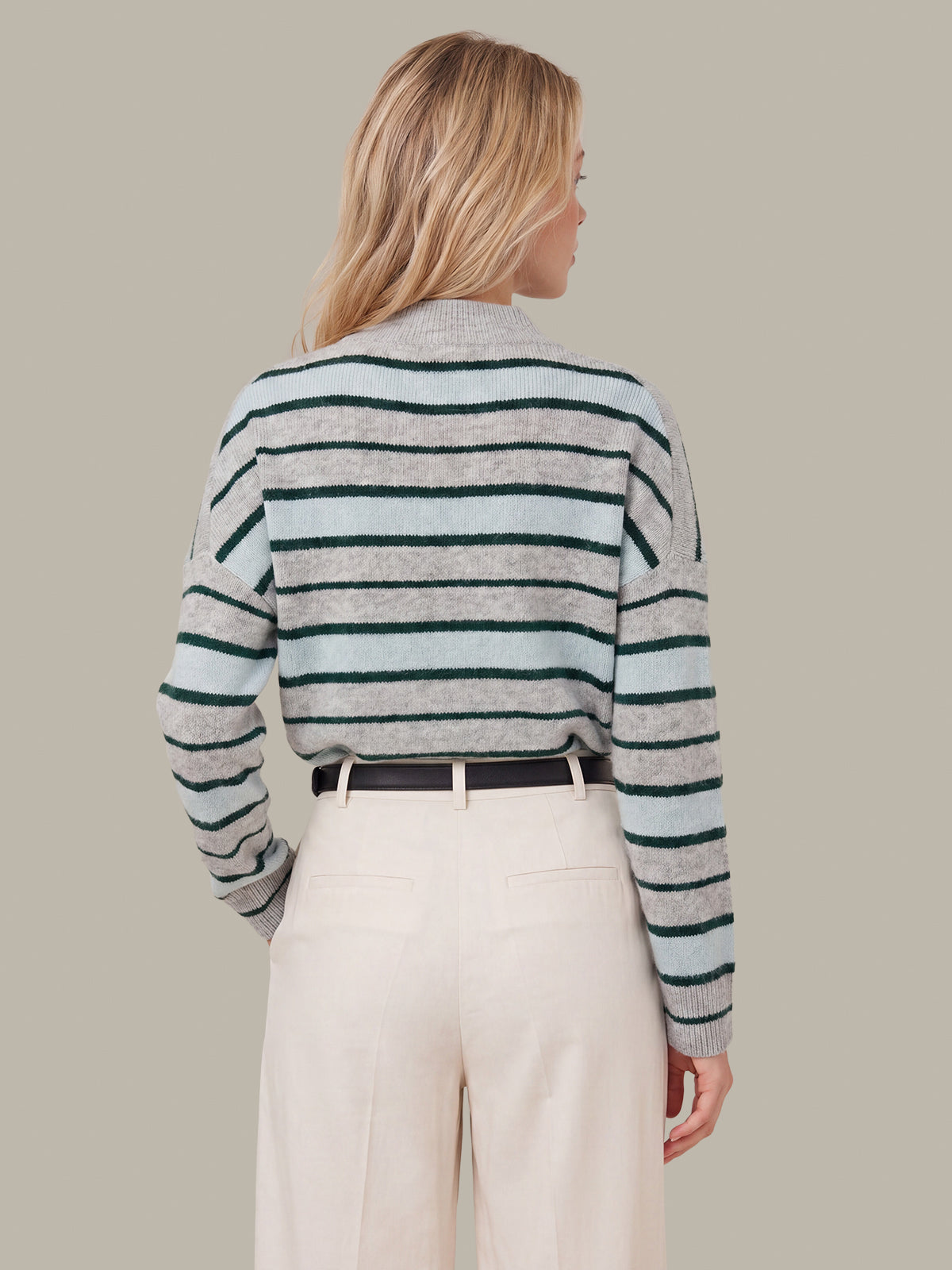 A woman shown from the back wearing a cropped, long-sleeve knit sweater with horizontal stripes in light gray and dark green, paired with cream-colored trousers and a black belt, against a solid beige background.