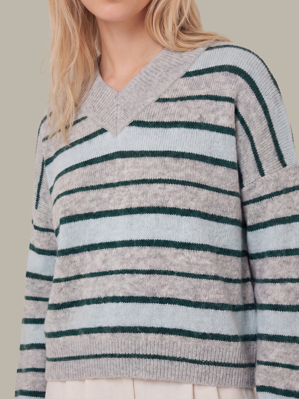 A close-up of a woman wearing a knit V-neck sweater with horizontal stripes in light gray and dark green, paired with a cream skirt against a neutral beige background.
