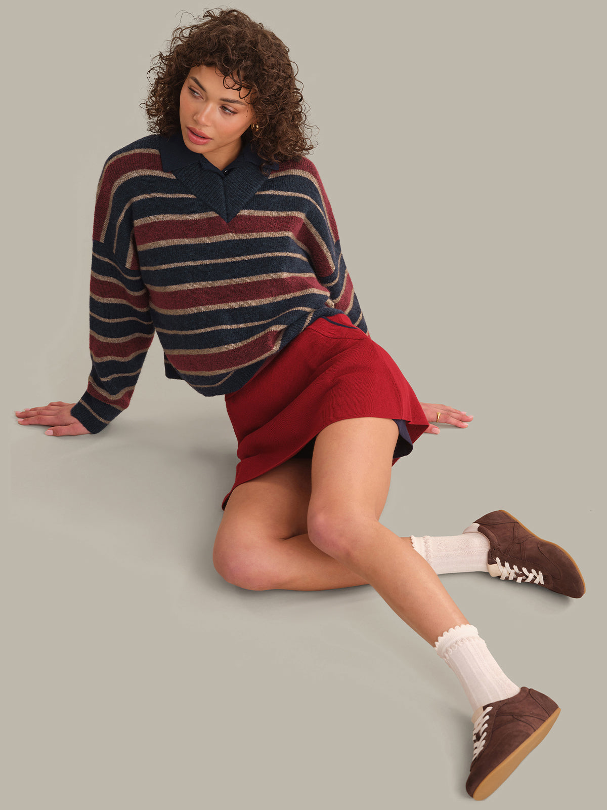 A model wearing a navy, burgundy, and beige striped knit polo sweater, a red mini skirt, white socks, and brown suede shoes, posing on a solid light gray studio background.