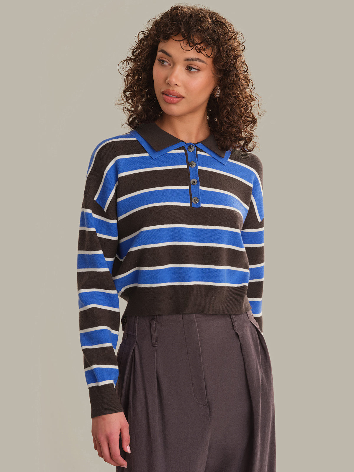 A woman with curly brown hair wearing a long-sleeve polo shirt with horizontal stripes in blue, brown, and white, paired with dark gray pleated trousers, against a solid light gray background.