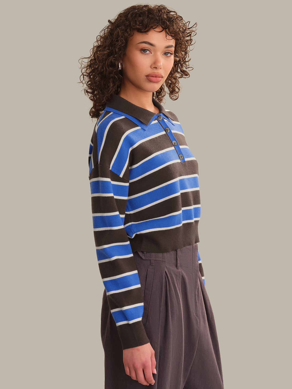 A woman wearing a long-sleeve polo shirt with horizontal stripes in blue, brown, and white, paired with dark gray trousers, set against a solid light gray background.