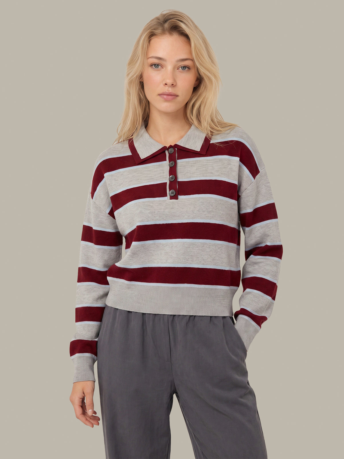 A woman modeling a long-sleeve knit polo sweater with horizontal gray and burgundy stripes, paired with gray pants, against a solid light gray background.