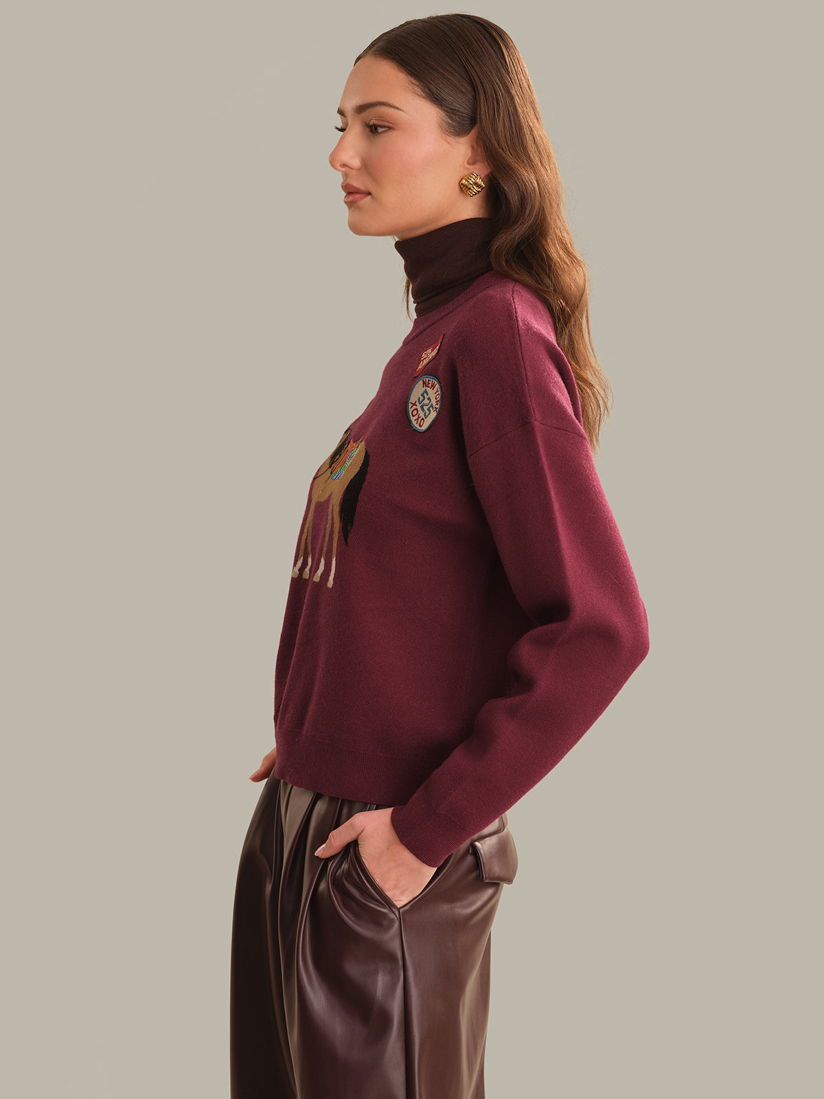 A woman in side profile wearing a burgundy crewneck sweater with a graphic print on the front and brown faux-leather pants, set against a solid gray studio background.