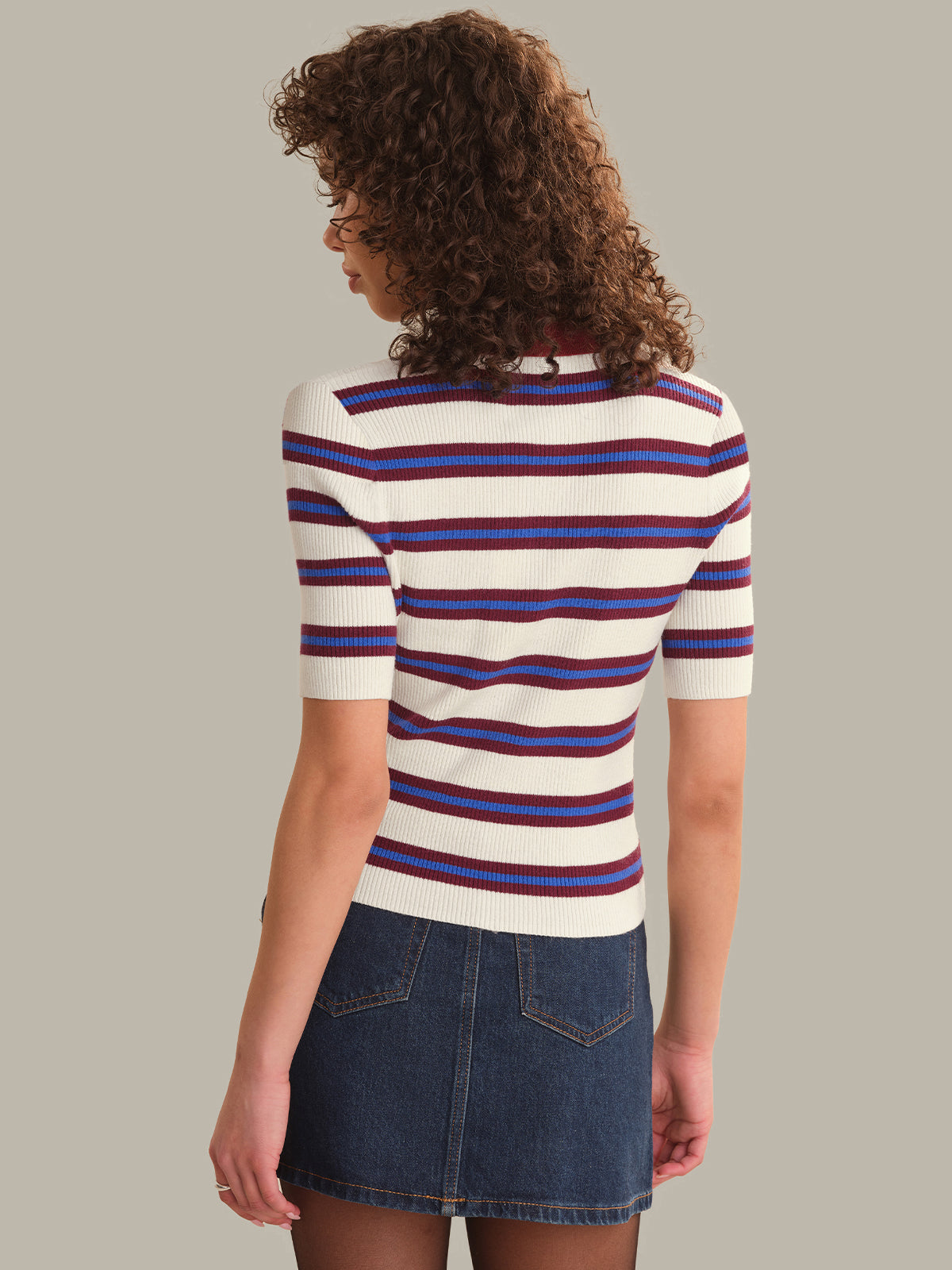 A woman shown from the back wearing a short-sleeve knit top with horizontal cream, red, and blue stripes, paired with a denim mini skirt, against a solid taupe background.