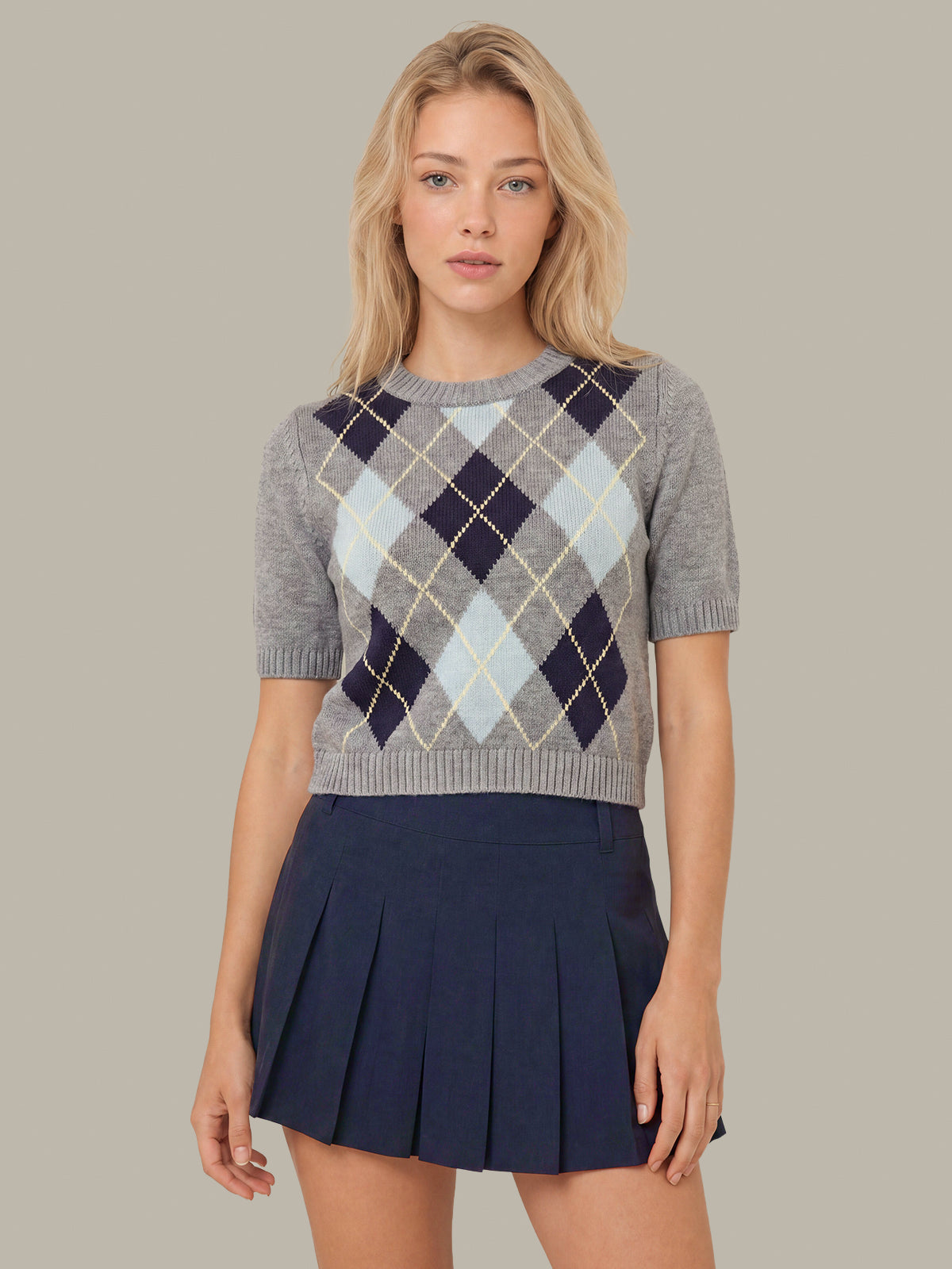 A woman modeling a short-sleeve knit sweater with an argyle pattern in gray, navy, light blue, and cream, paired with a navy pleated mini skirt, against a solid taupe background.