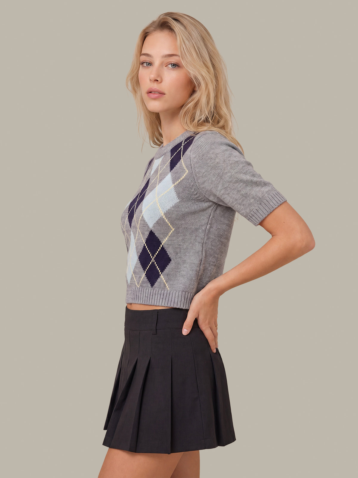 A woman modeling a gray short-sleeve cropped knit top with an argyle pattern in navy, light blue, and beige, paired with a dark pleated mini skirt, against a solid beige background.