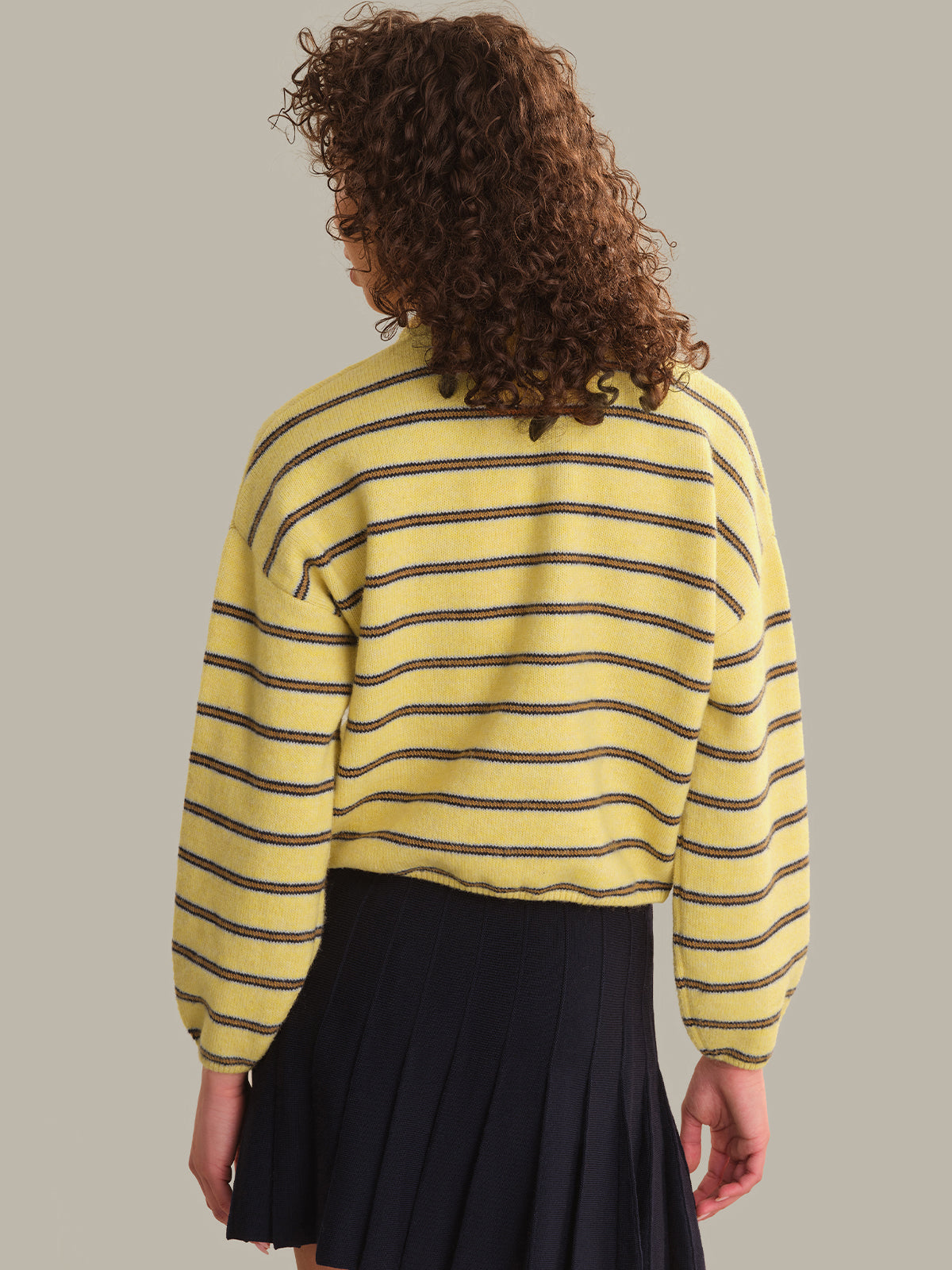 A person shown from the back wearing a yellow knit sweater with thin dark horizontal stripes and a dark pleated skirt, against a plain warm gray studio background.