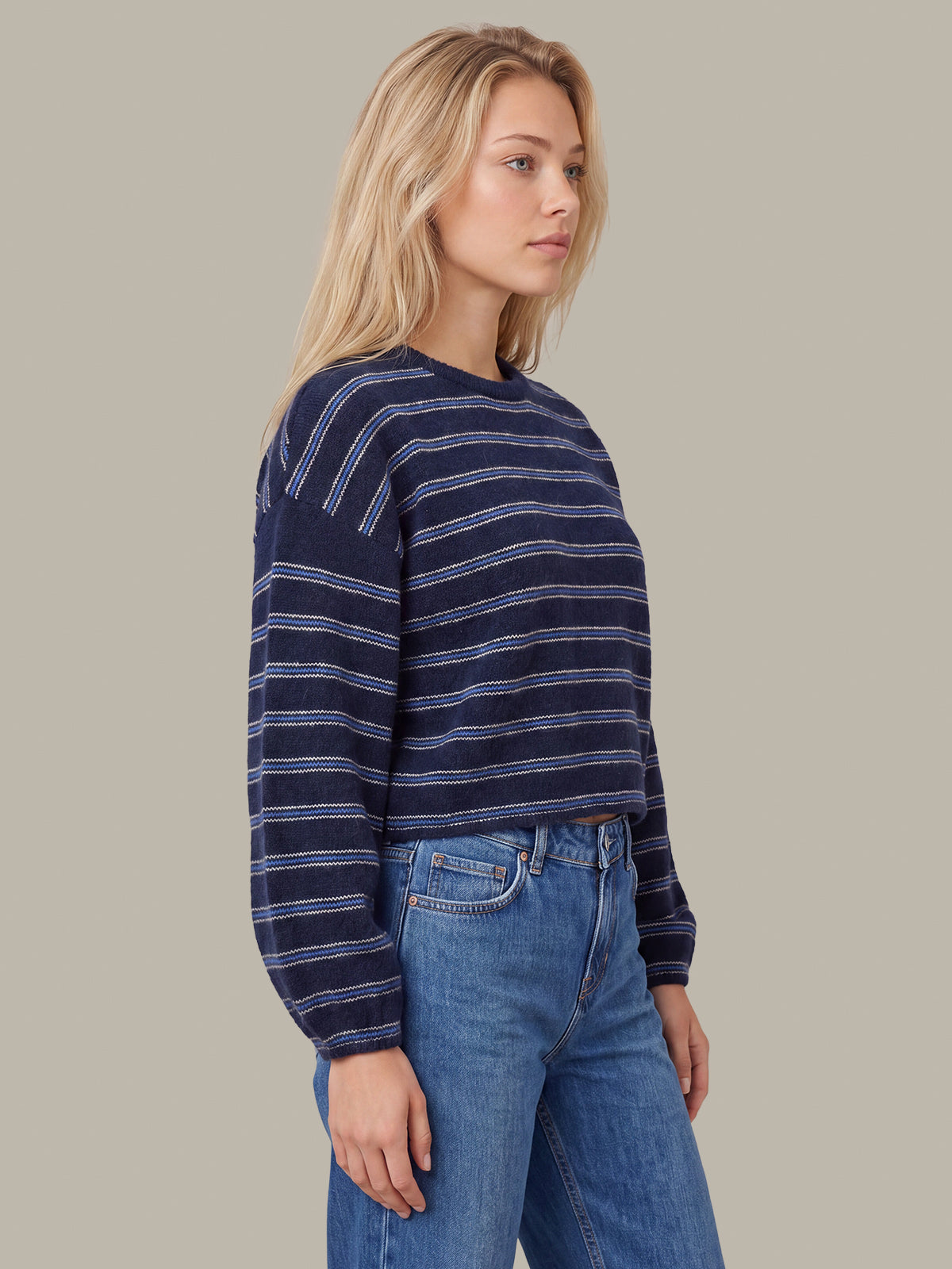 A woman wearing a navy blue and white striped long-sleeve knit top with blue jeans, shown from the waist up against a solid beige background.