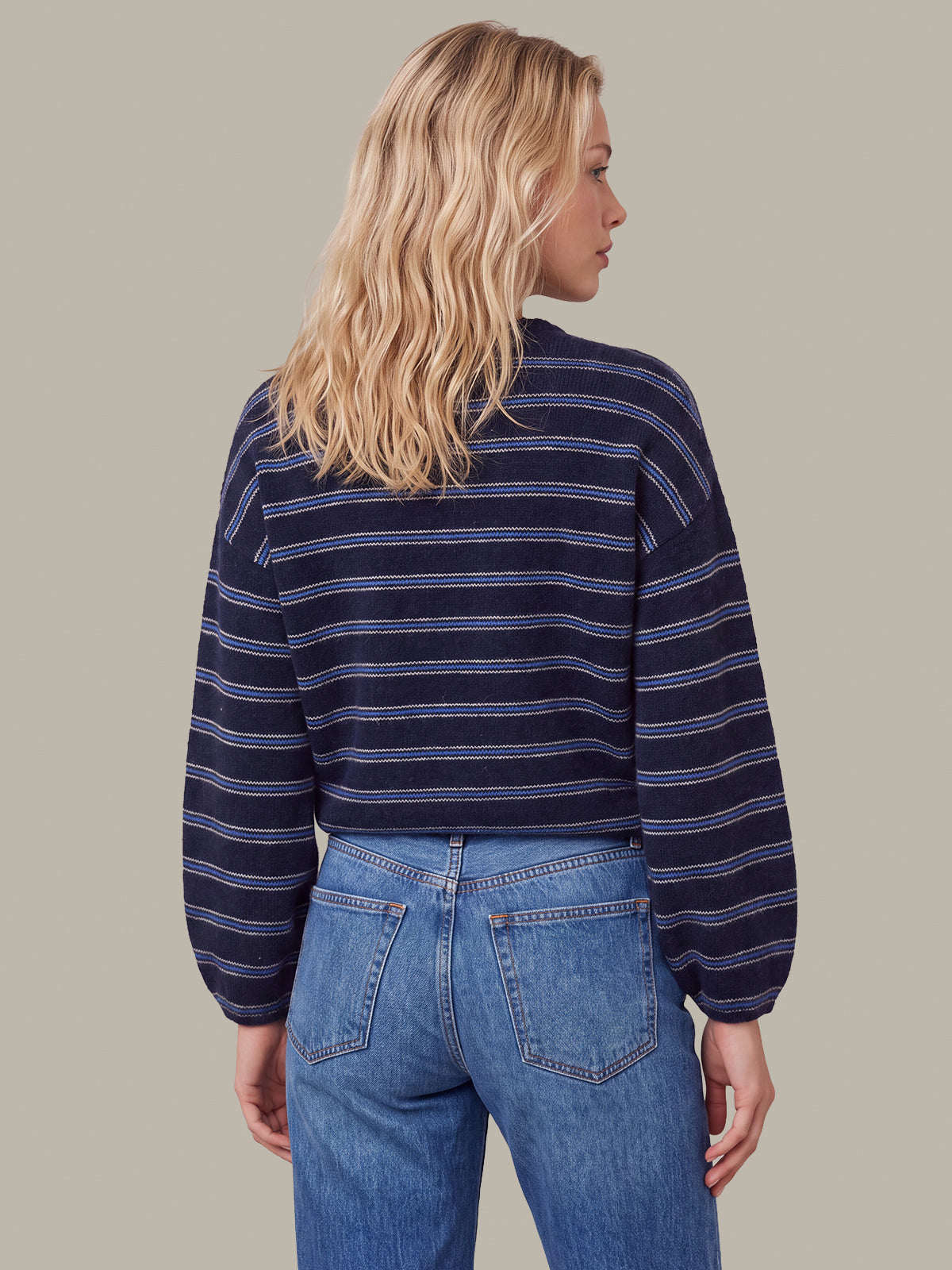 A woman shown from the back wearing a navy blue long-sleeve cropped sweater with thin horizontal light blue stripes, paired with blue jeans, against a solid light gray background.