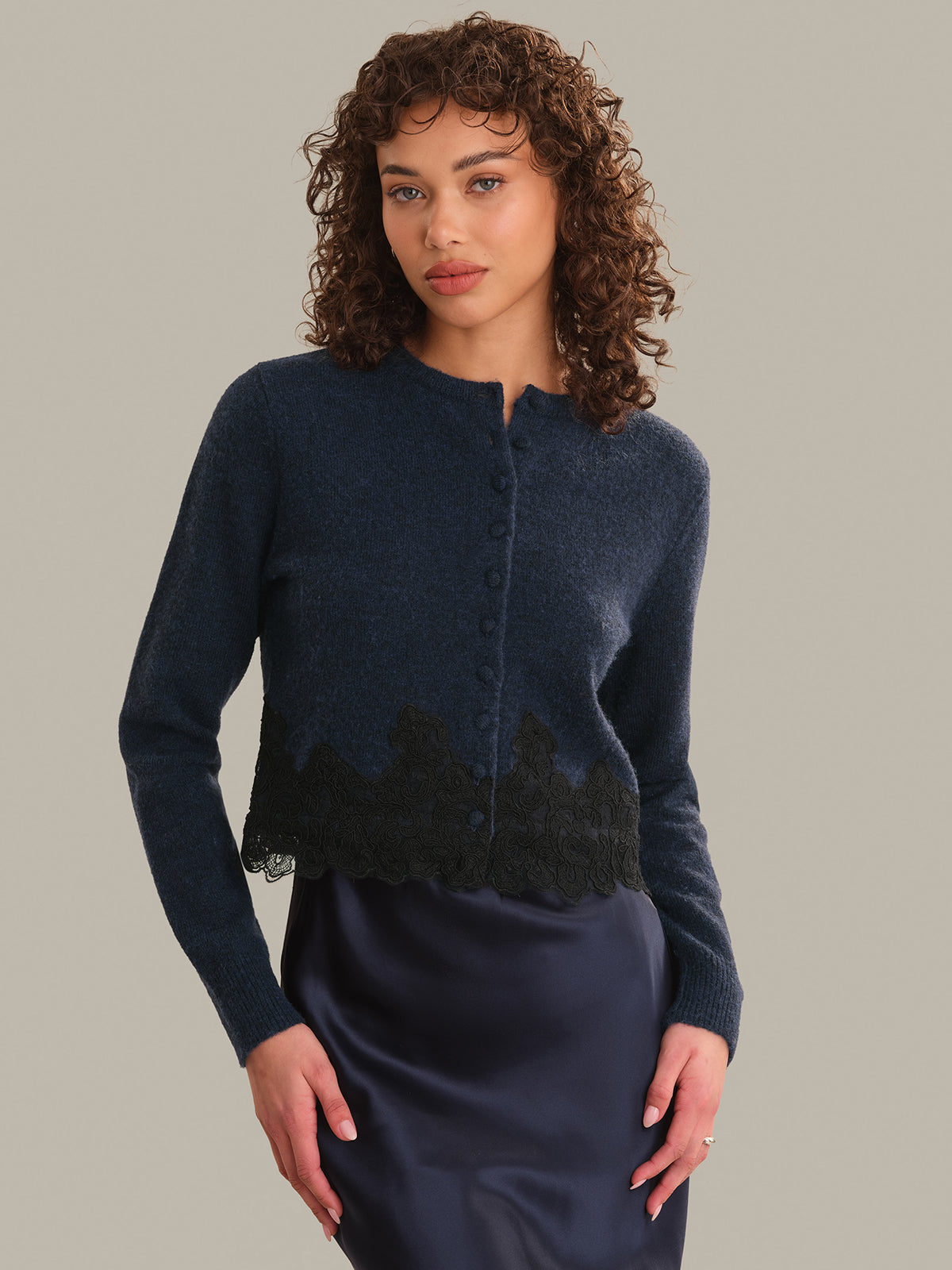 A woman with curly brown hair models a navy blue knit cardigan featuring a black lace hem, paired with a matching dark navy skirt, against a solid light gray background.