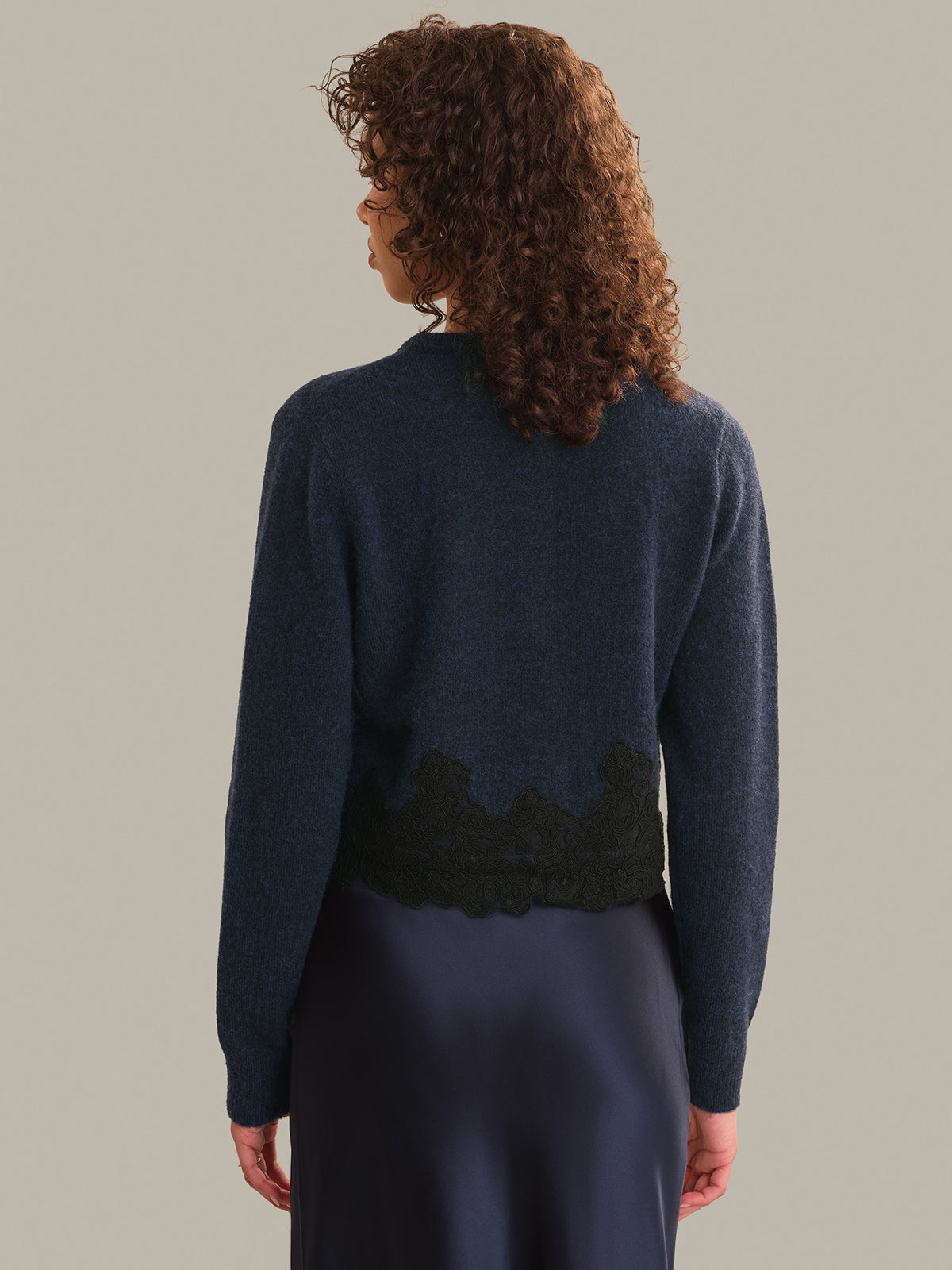 A woman shown from the back wearing a navy blue knit cardigan with a black lace hem detail, paired with a dark navy skirt, against a solid light gray background.