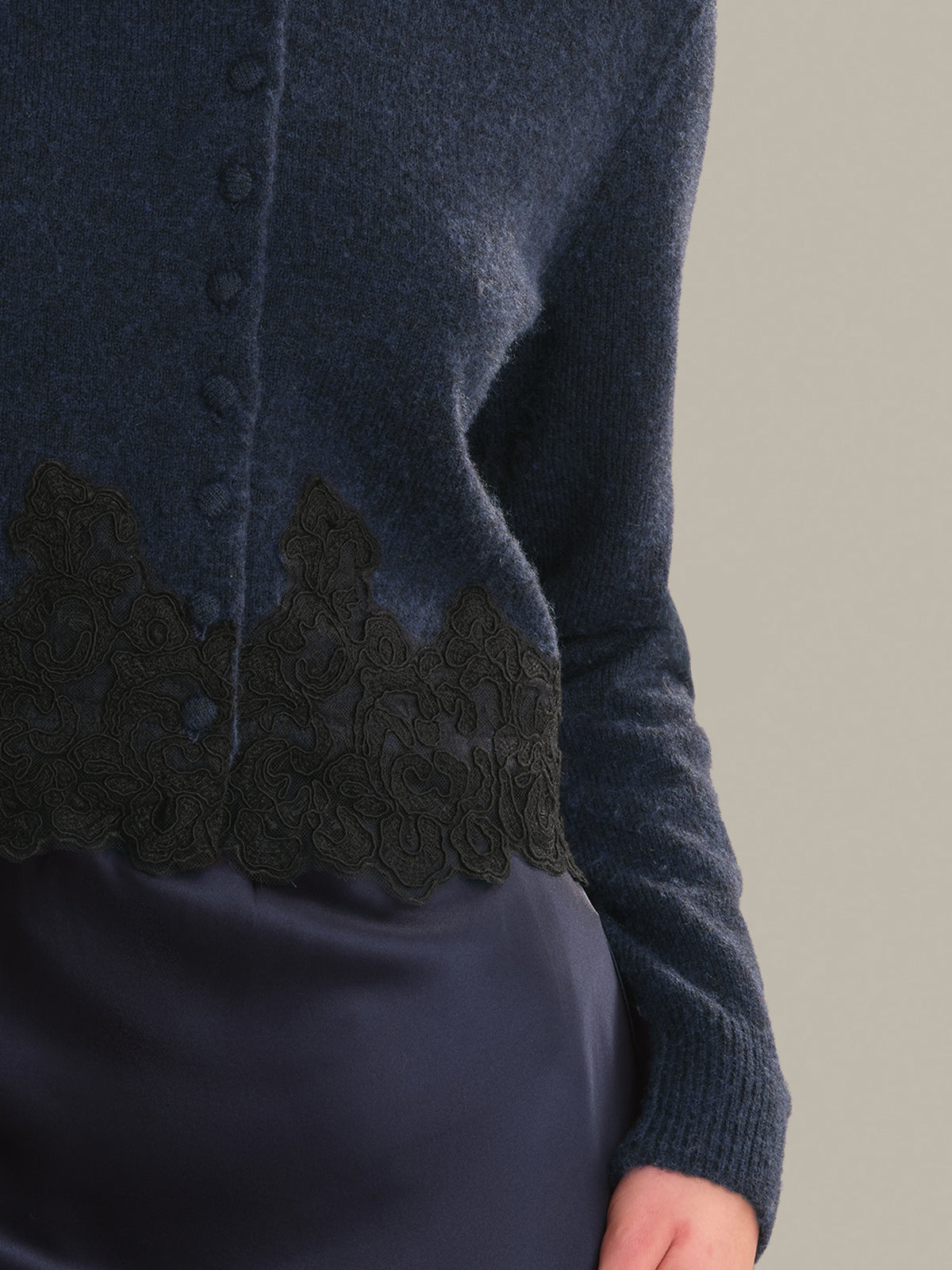 Close-up of a navy blue knit cardigan with black lace trim along the button placket, worn by a model against a plain beige background.