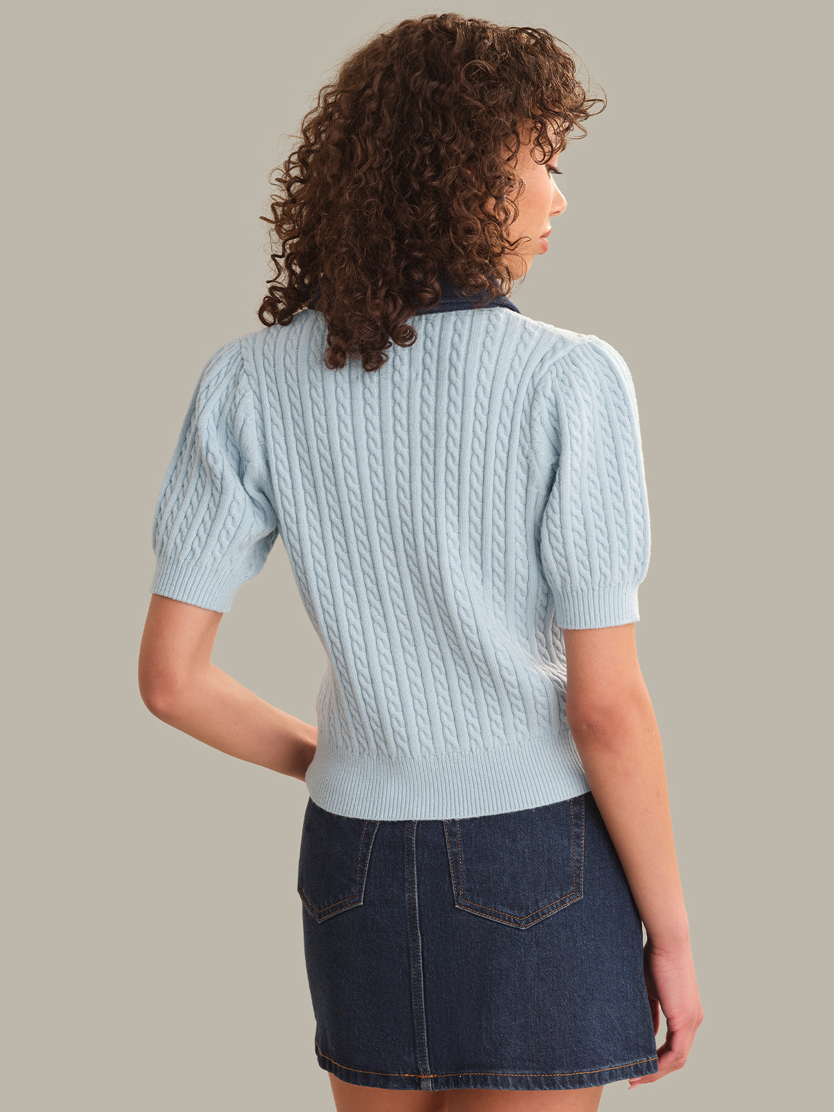 Woman wearing a light blue cable knit sweater and dark blue denim skirt on a neutral background