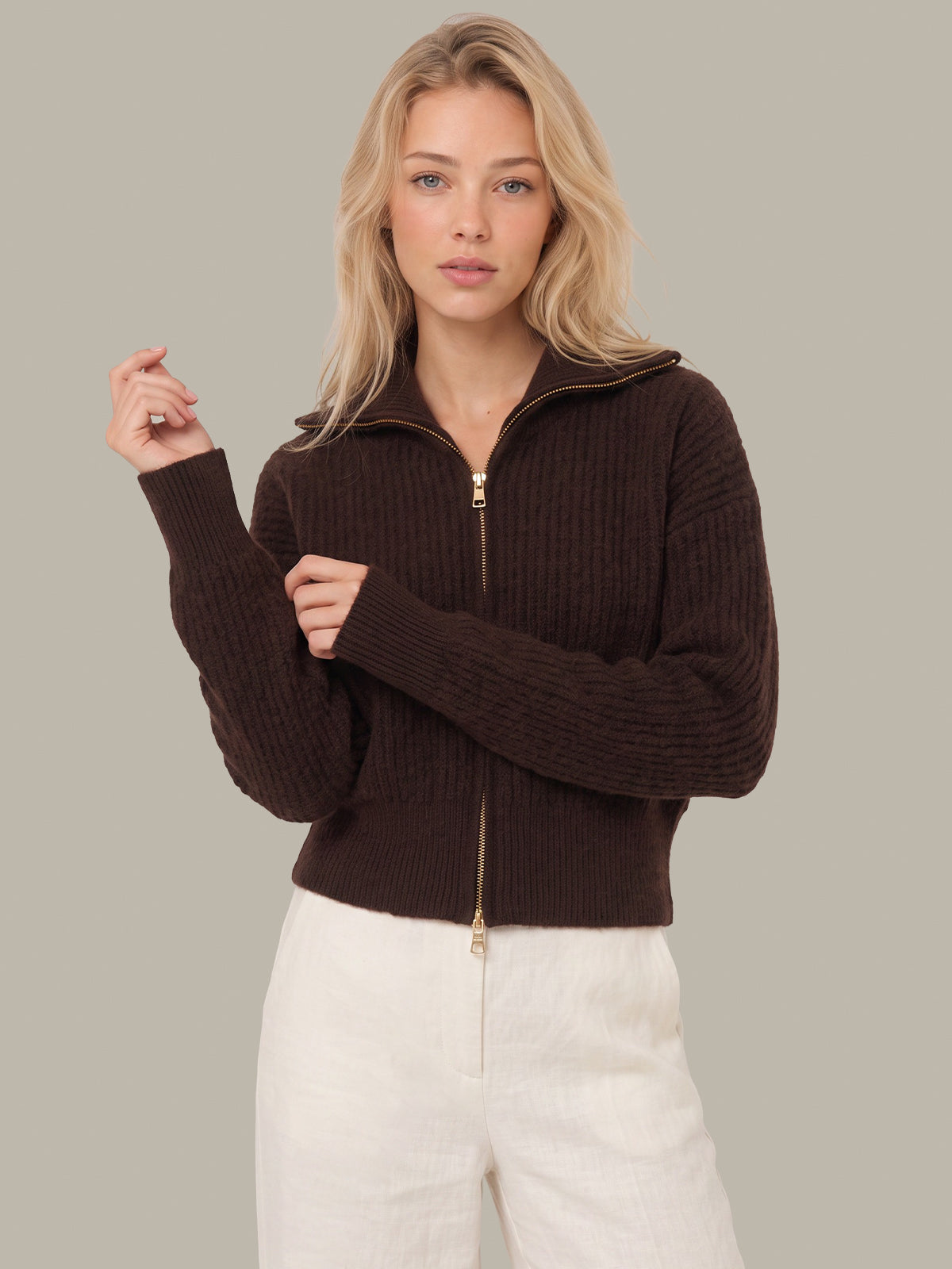 A blonde woman wearing a dark brown ribbed knit zip-up sweater with a stand collar and white pants, posed against a solid light gray studio background.