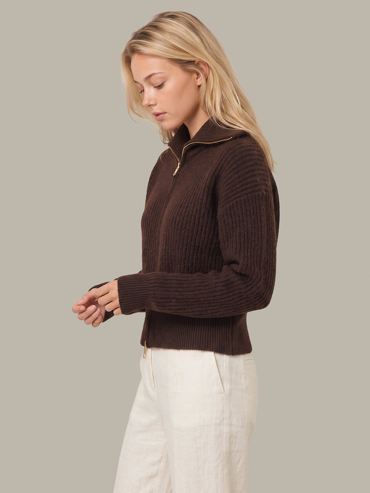 A woman wearing a dark brown ribbed knit half-zip sweater with gold trim and white trousers, shown from the waist up against a solid light gray background.