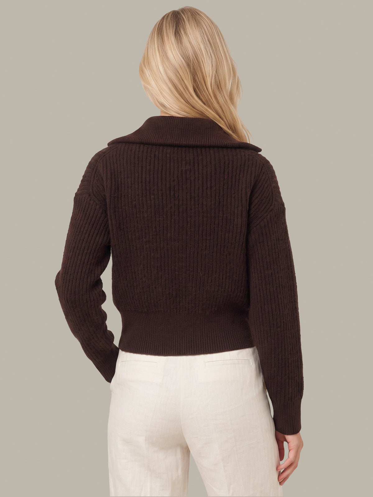 A woman shown from the back wearing a dark brown ribbed knit sweater with a wide collar and cream pants, set against a solid light gray background.