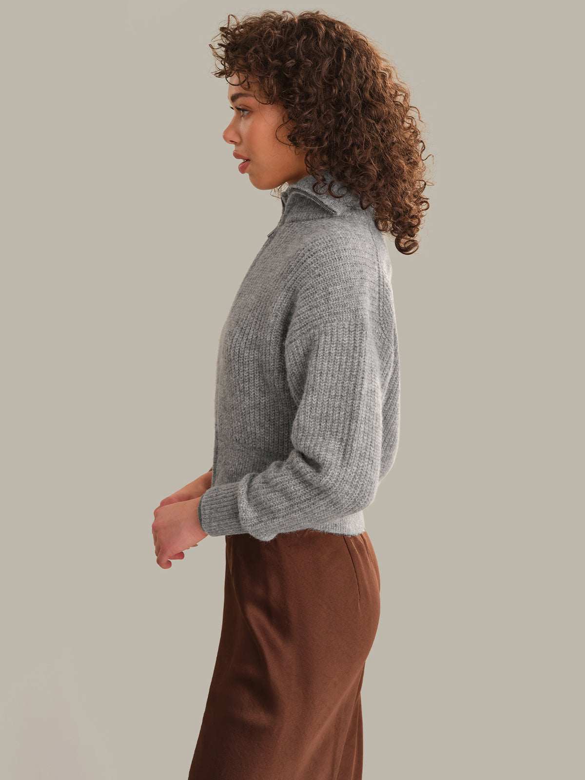 A woman with curly brown hair is shown in profile wearing a gray ribbed mock-neck sweater and a brown skirt, set against a solid light gray background.