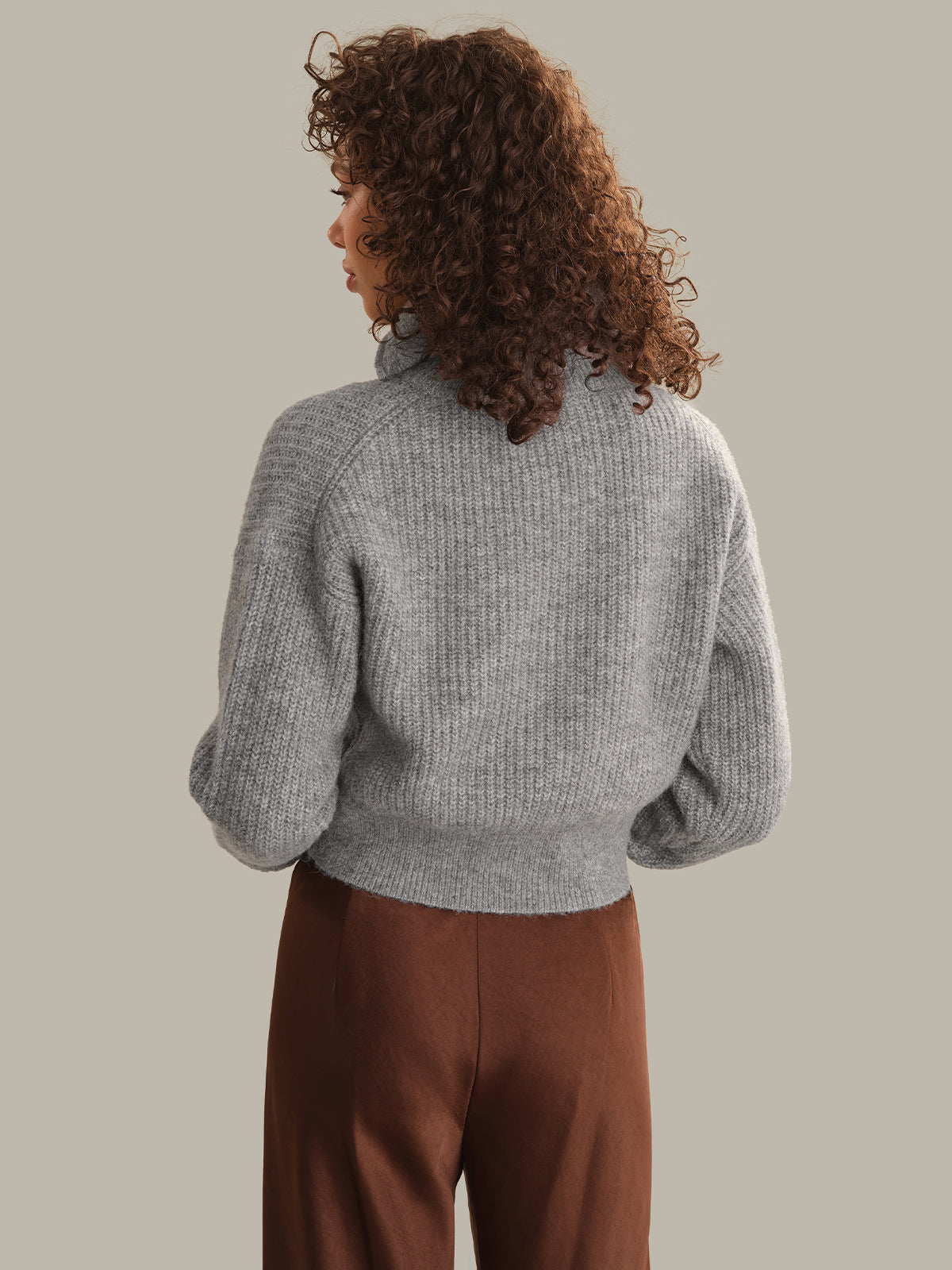 A woman with curly brown hair is shown from behind wearing a light gray ribbed knit mock-neck sweater and brown pants, set against a solid warm gray background.