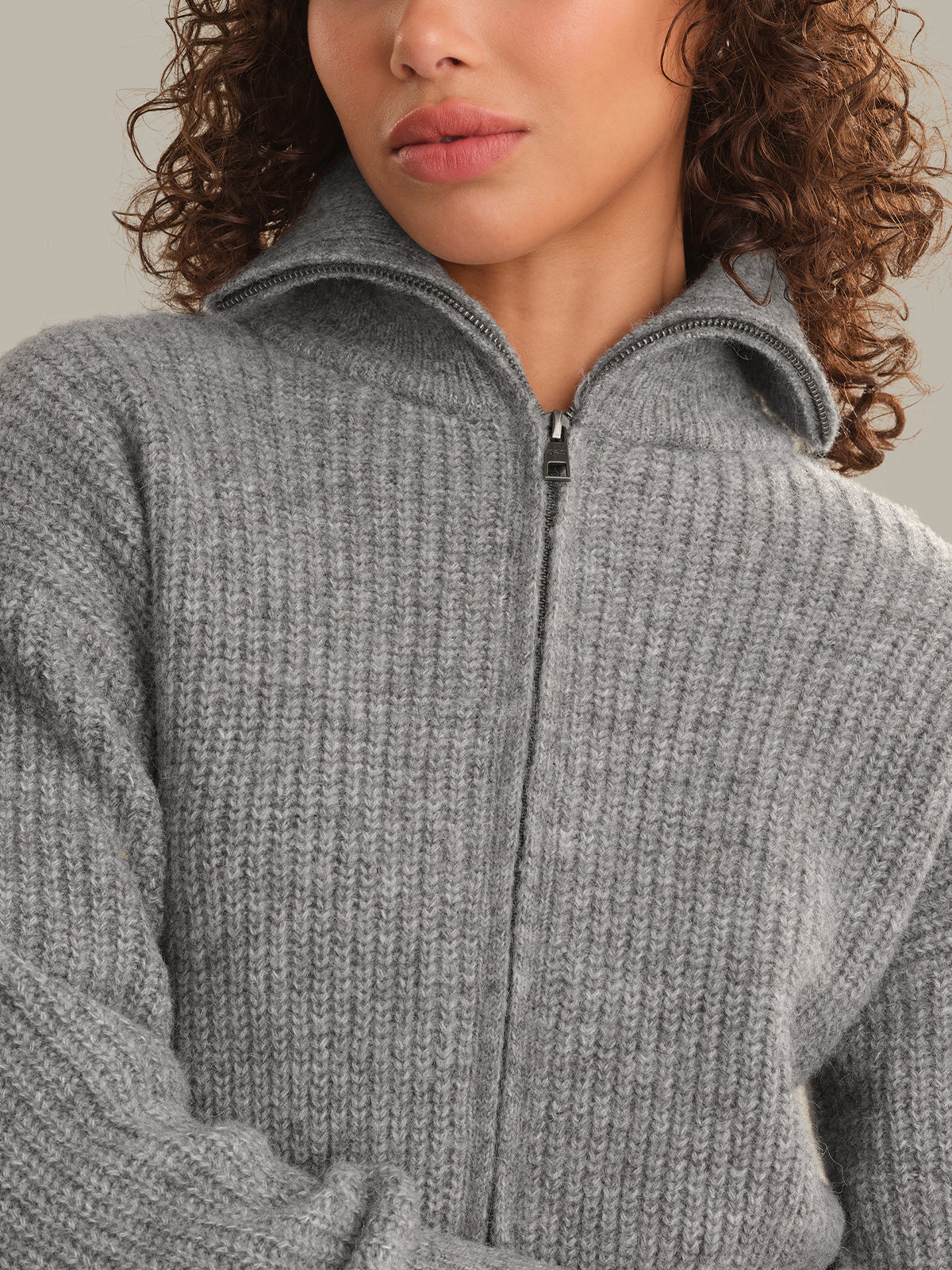 Close-up of a woman wearing a gray ribbed knit zip-up sweater with a stand collar, cropped from the lower face to the torso against a plain light gray background.