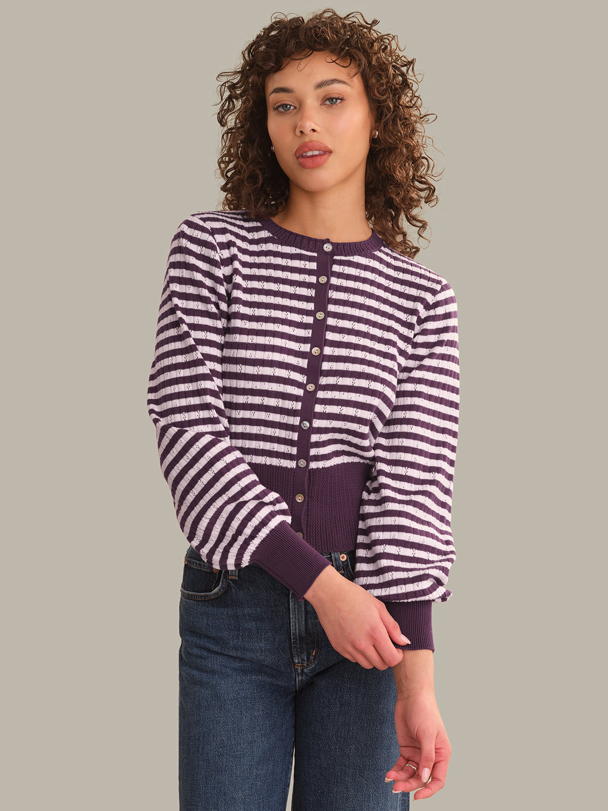 A woman with curly brown hair wearing a purple and white striped long-sleeve button-up cardigan with dark purple ribbed trim, paired with dark blue jeans, against a solid light gray background.