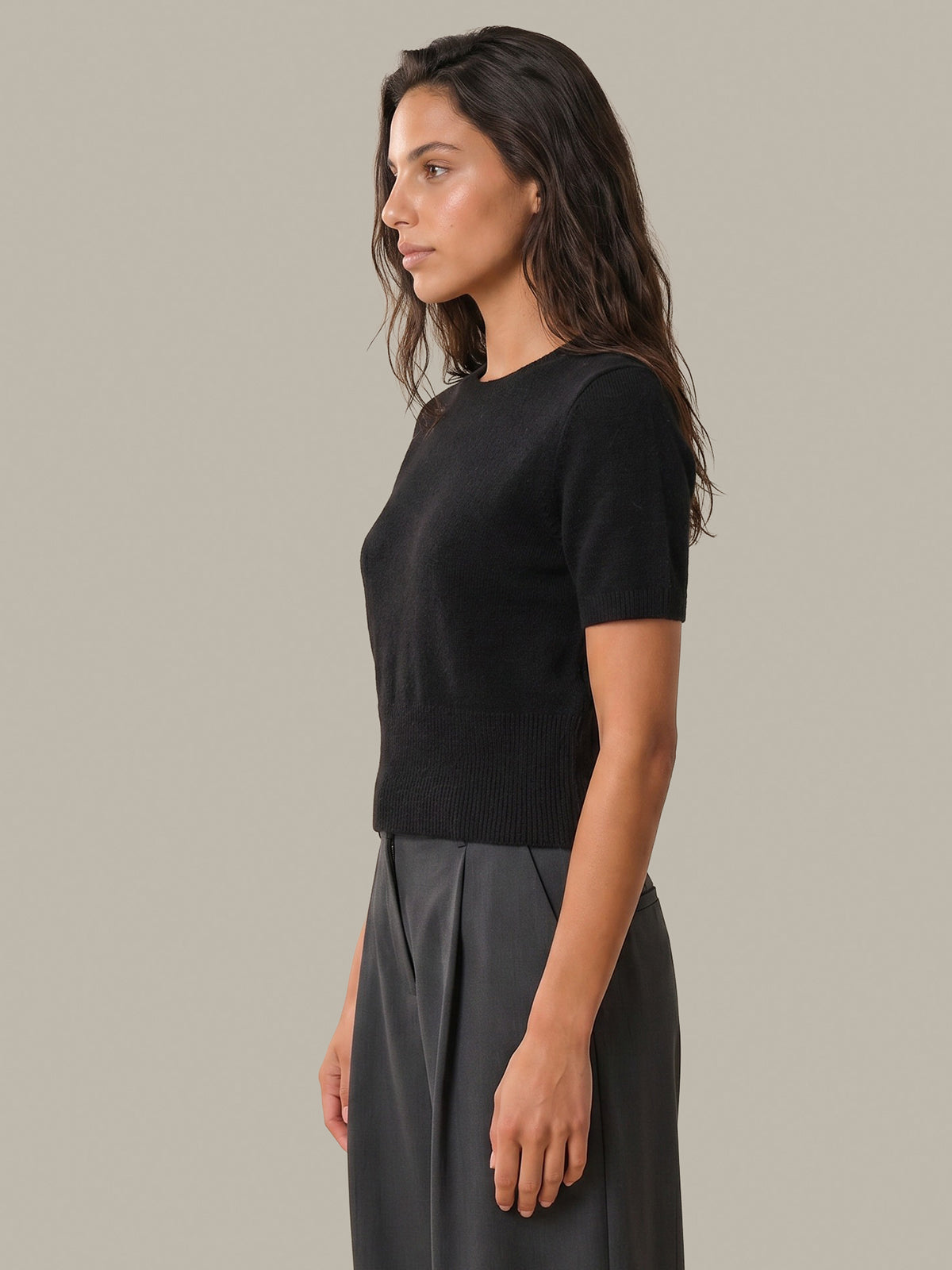 A woman shown in side profile wearing a fitted black short-sleeve knit top and dark gray pleated trousers, photographed against a plain warm gray studio background.