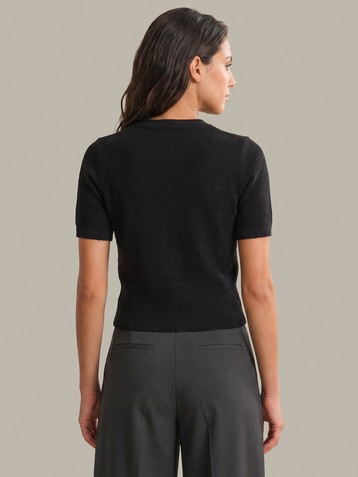 A woman shown from the back wearing a short-sleeve black knit top and dark gray wide-leg trousers, photographed against a plain warm gray studio background.