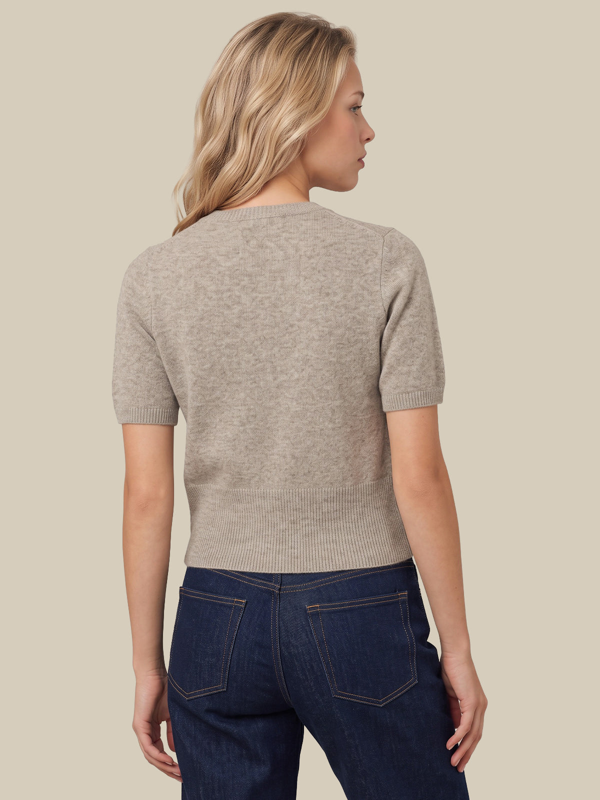 A woman shown from the back wearing a short-sleeve ribbed knit sweater in a light taupe-beige color, paired with dark blue jeans, against a solid light beige background.