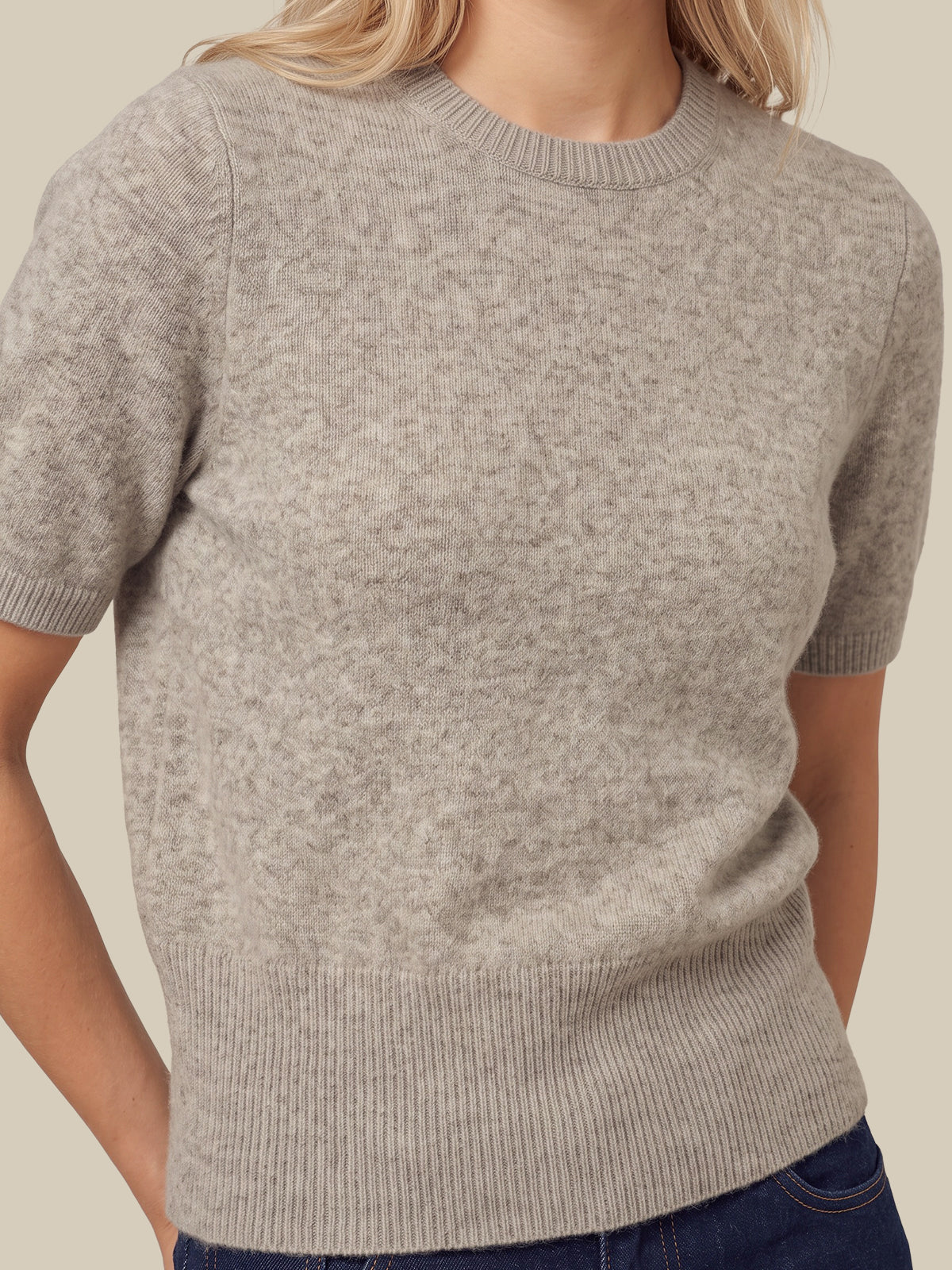 A close-up of a woman wearing a short-sleeve crewneck knit sweater in a light heather gray color, paired with dark blue jeans against a solid beige background.
