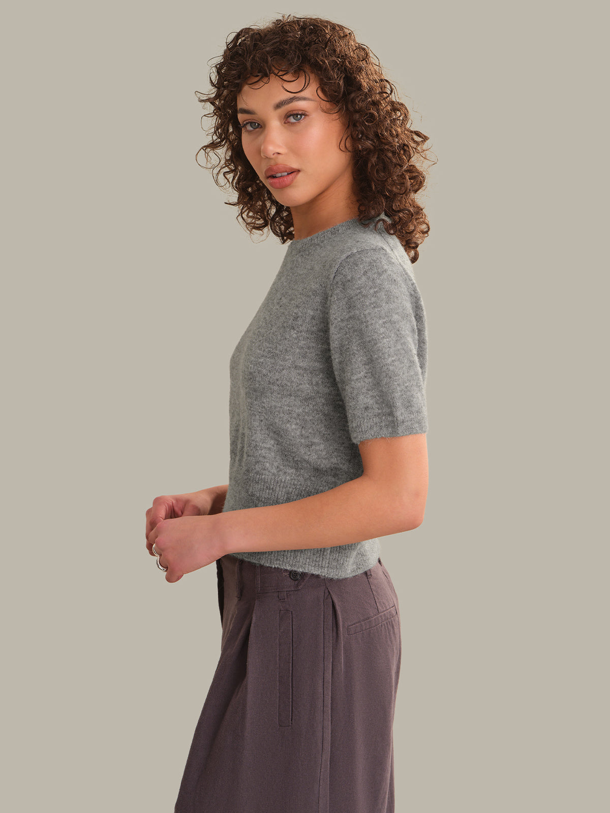 A woman with curly brown hair is shown in profile wearing a short-sleeve heather gray knit top and dark gray wide-leg trousers, set against a solid light gray background.
